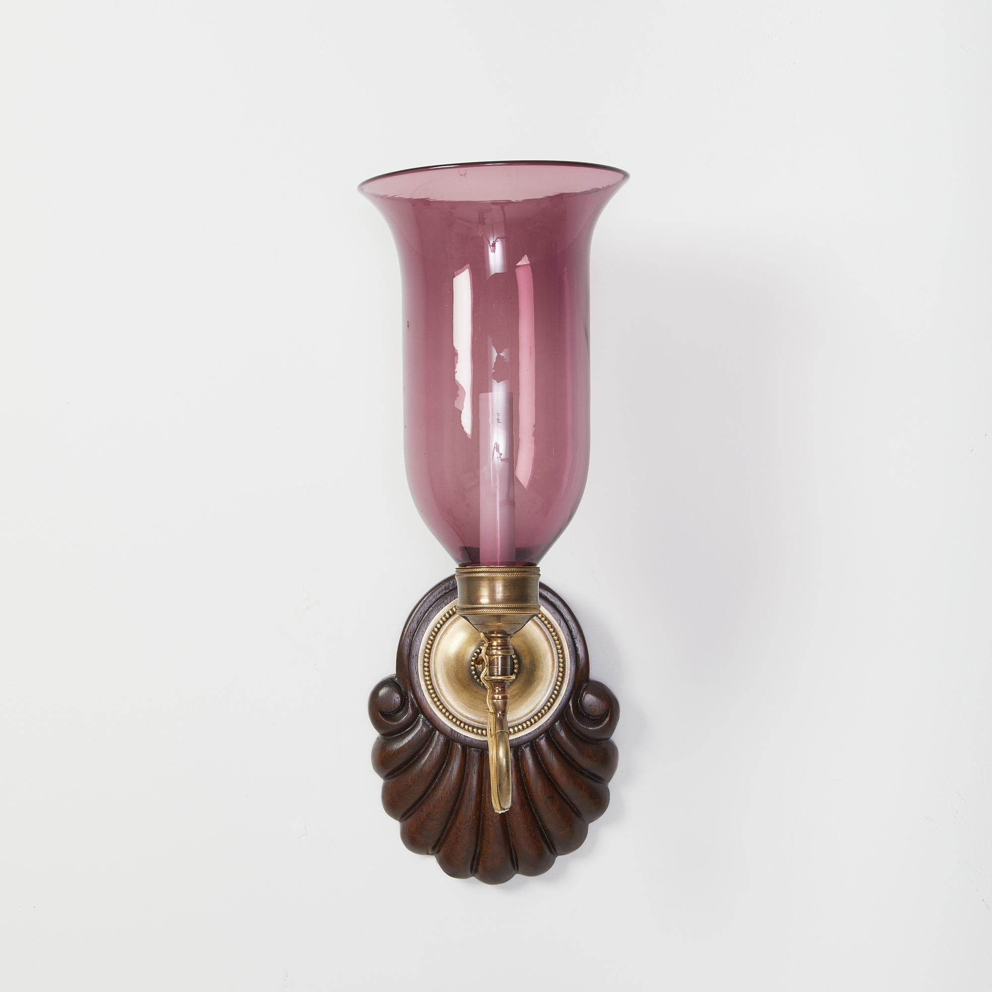 A pair of antique hand-carved shell form mahogany Anglo Indian styled backplates with patinated brass sconce fittings and purple glass flared hurricane shades. Electrified with one candelabra base socket per sconce.

Our custom hurricane sconces are