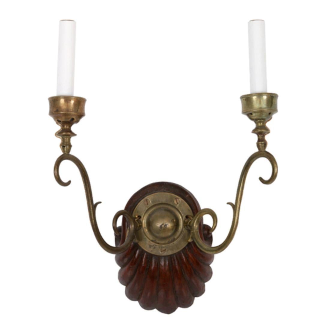 Cette paire d'appliques à double bras en bois et en laiton d'origine anglo-indienne présente une plaque arrière en bois sculpté avec un motif de coquillage, complétant deux bras courbés en laiton qui s'étendent vers l'extérieur. Les accoudoirs sont