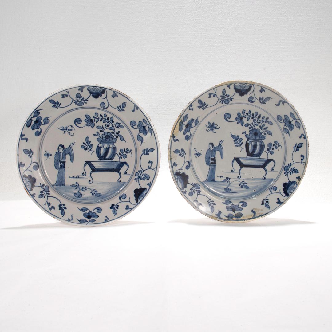 A pair of fine antique Dutch Delft plates.

Each depicting a woman in classical Chinese garb gesturing towards an extravagant potted plant atop a table.

The rim of each is decorated with floral accents and a geometric border.

Simply a wonderful