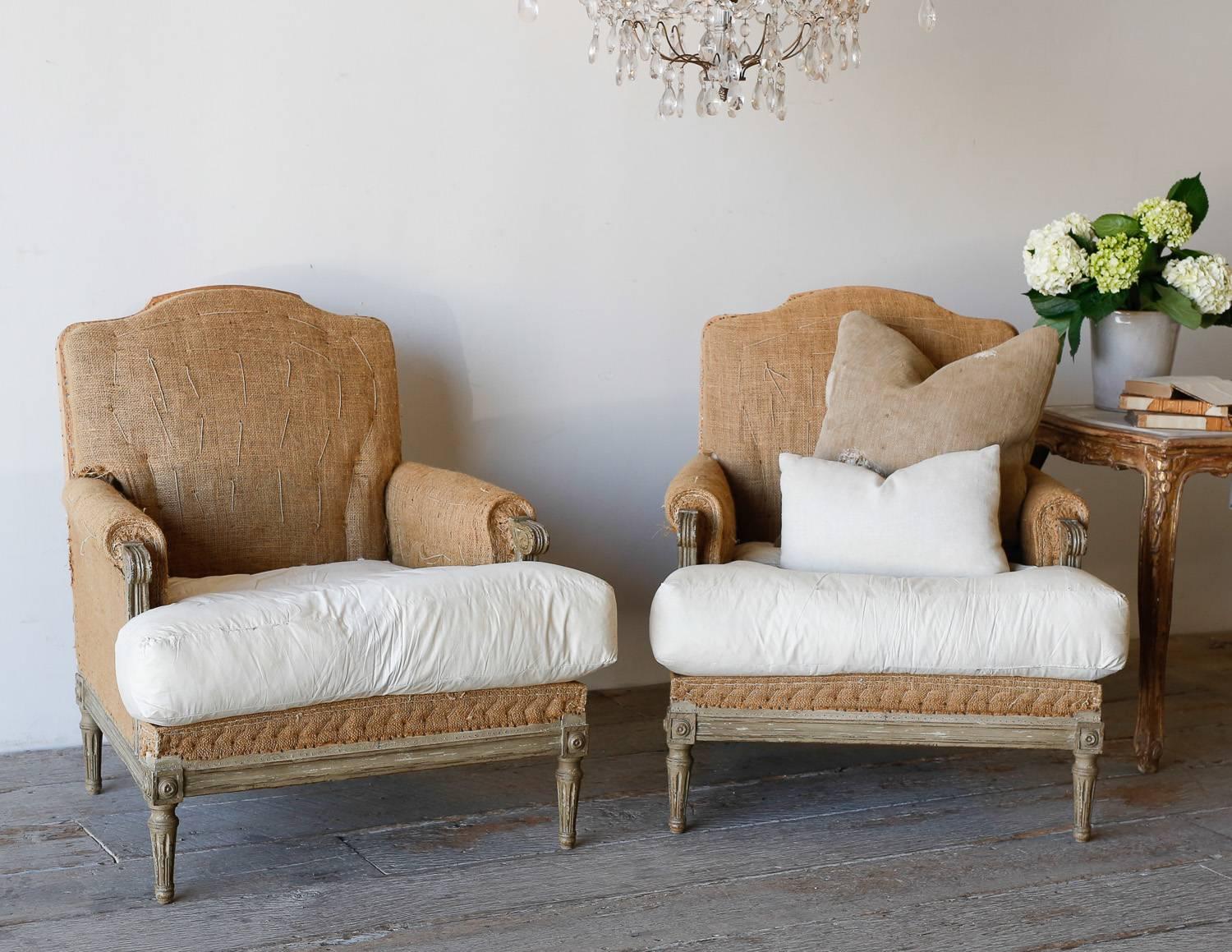 Stunning pair of bergeres in sandy olive. The chairs boast fluffy, comfortable down cushions and deep proportions. Upholstered in burlap with exposed wood backs and Classic Louis XVI fluted legs. Carved medallions decorate the legs and make this