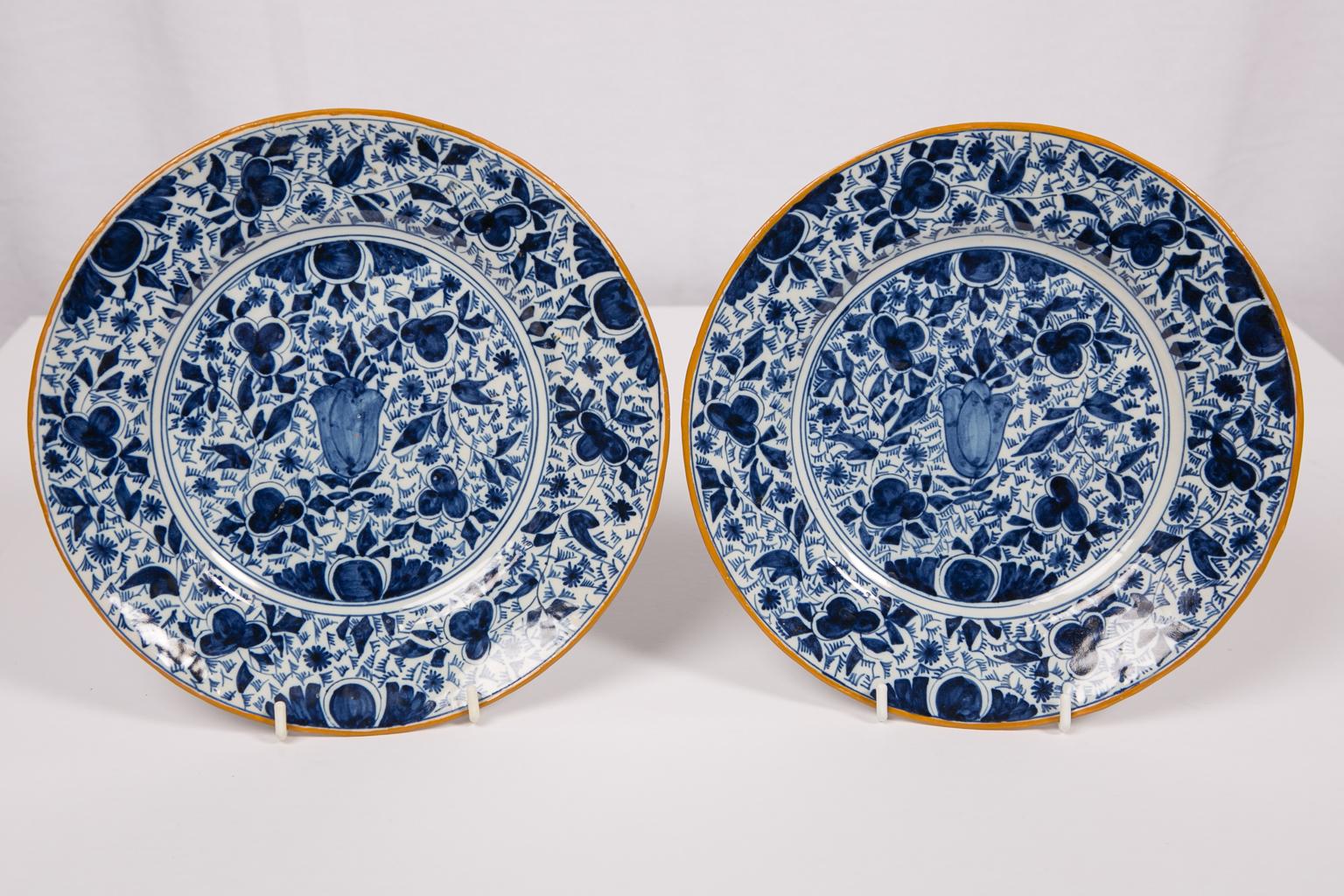 We are pleased to offer this pair of antique Delft blue and white plates which are hand-painted with a central tulip bulb and an overall pattern of flowers and leaves. The edge is painted with ochre colored slip. The ochre colored edge brings life