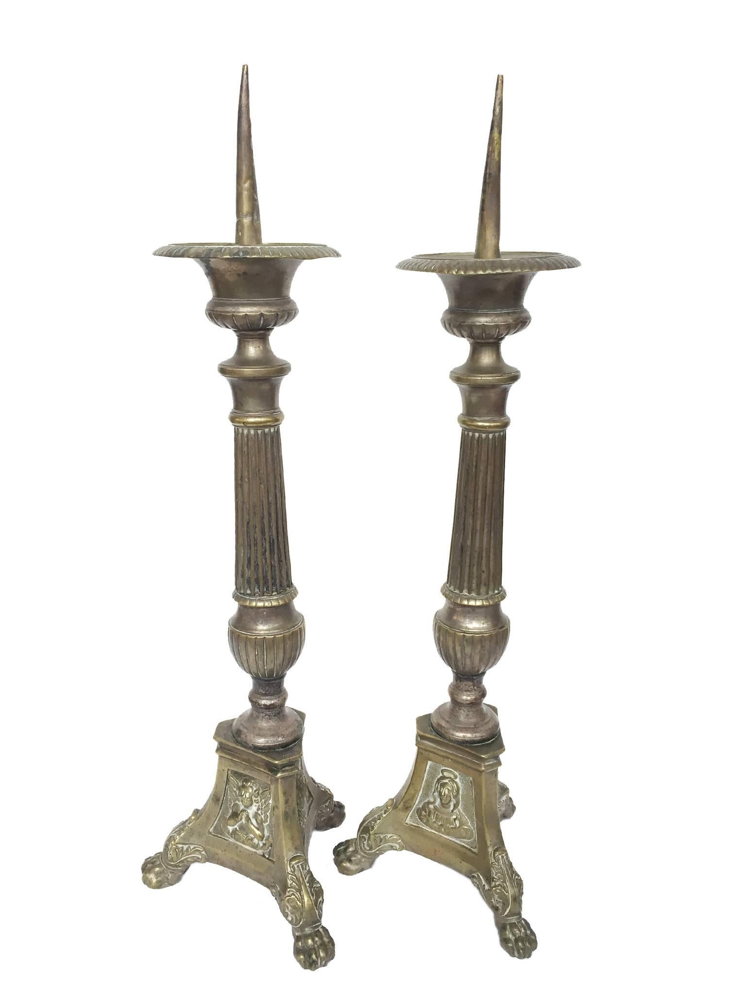 These two candleholders are comprised of brass with molded details. These include fluted stems with a triangular base that end in lion's feet. The three sides of the base bear an embossed image of Jesus Christ, the Virgin Mary, and a cherubic angel.