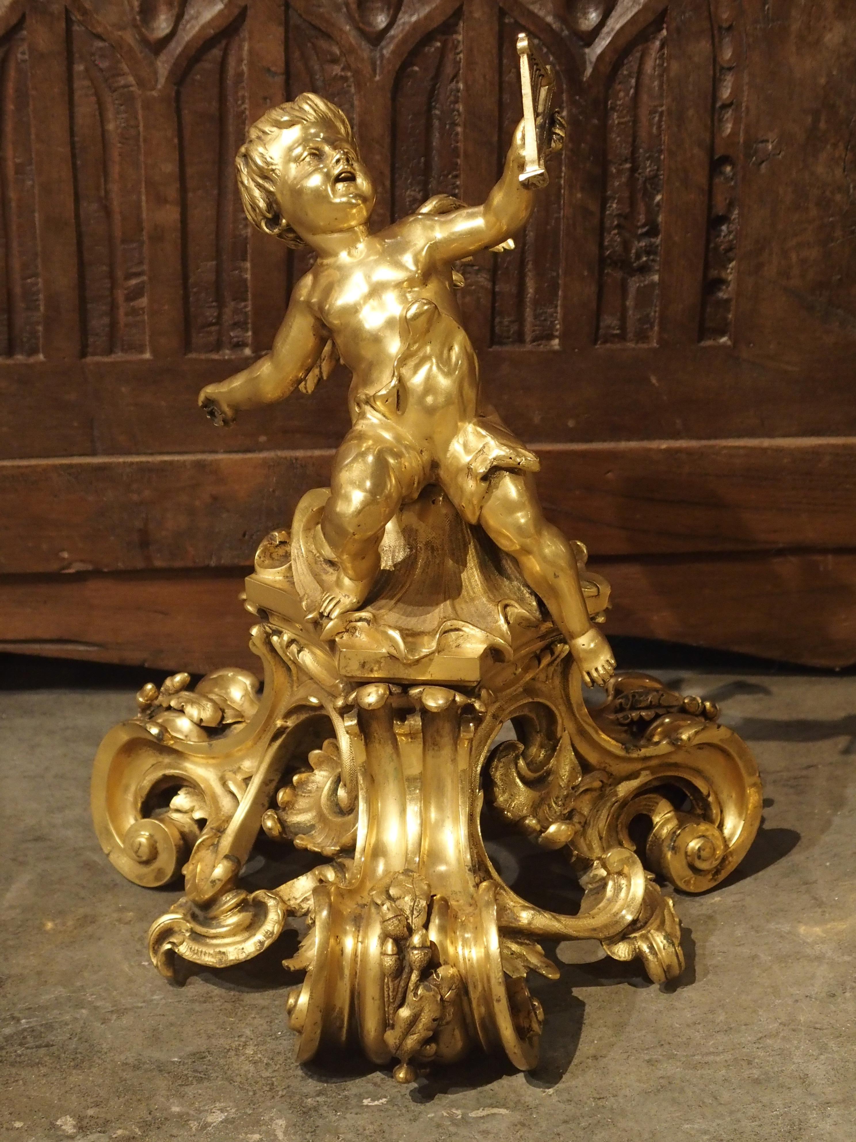 This is an outstanding pair of antique French gilt bronze chenets from the mid to late 1800s. Two cherubs rest upon decadent platforms composed of 3 thick C-Scrolled legs adorned with oak leaves, acorns, and shells. One of the cherubs has a