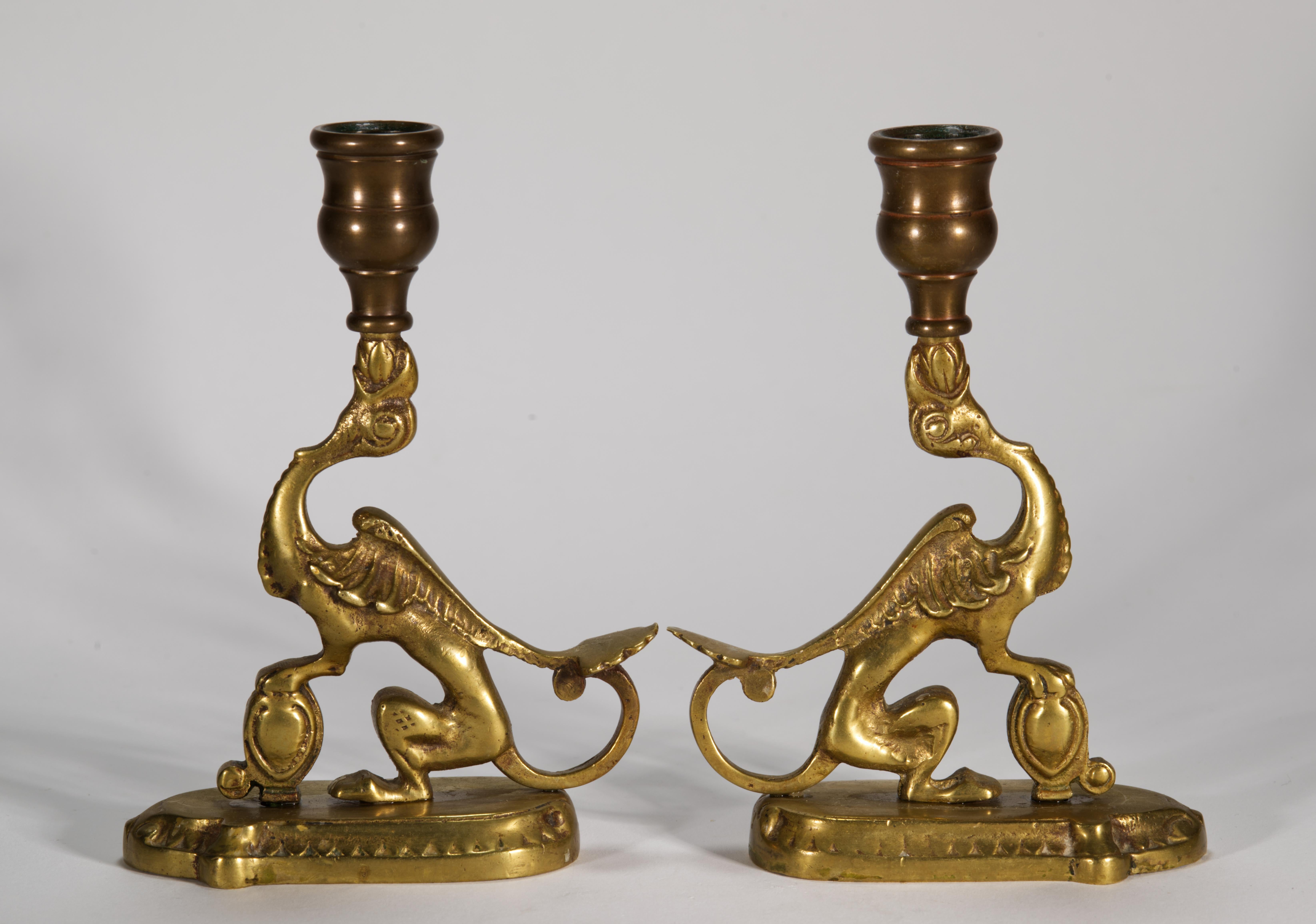  Pair of heavy bronze candle holders or chambersticks feature sitting griffons, holding the sconces with their beaks. The griffons are sitting on highly detailed elongated bases with their front paws resting on small oval vessels or shields; their