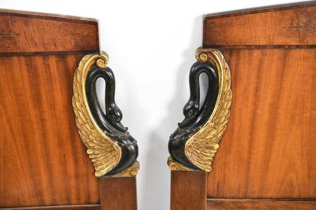 The pair of antique carved single headboards are in the Empire style. The carved black swans on either side have gilt wings, and painted cartouches are in the center. Condition: Loss to paint. Scratches, scuffs, wear consistent with age and use.
 