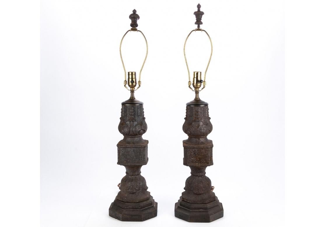 A large and very solid feeling pair of antique iron architectural elements as lamp bases. Three part shaped iron elements with decorative motifs with leaves on the tops and bottoms. On octagonal bases. Turned and painted wood urn form finials.
