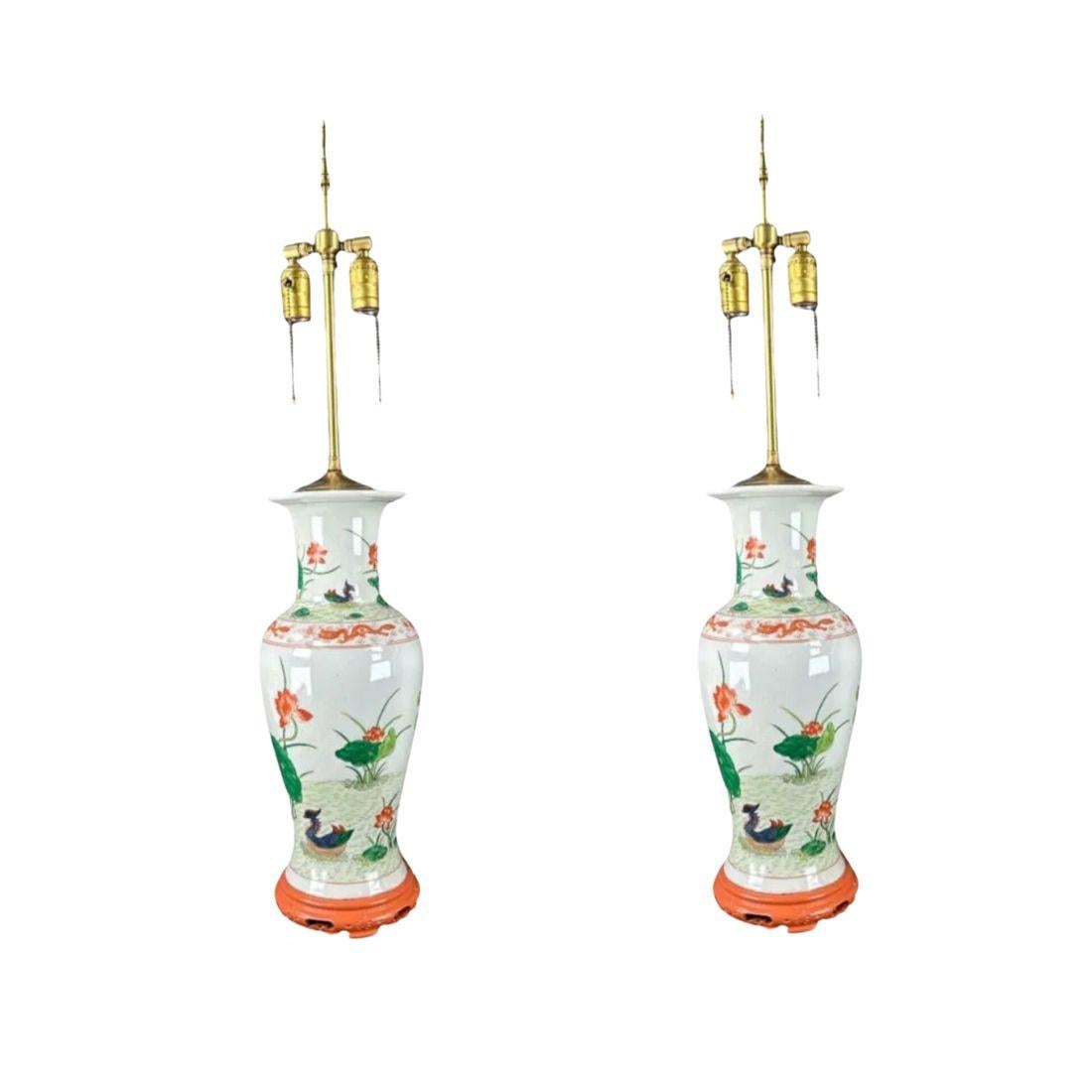 This elegant pair of antique Chinese porcelain lamps showcases intricate botanical and floral motifs hand-painted on a white base, accented with vibrant orange and green hues. Each lamp is mounted on a carved wooden base and complemented with a
