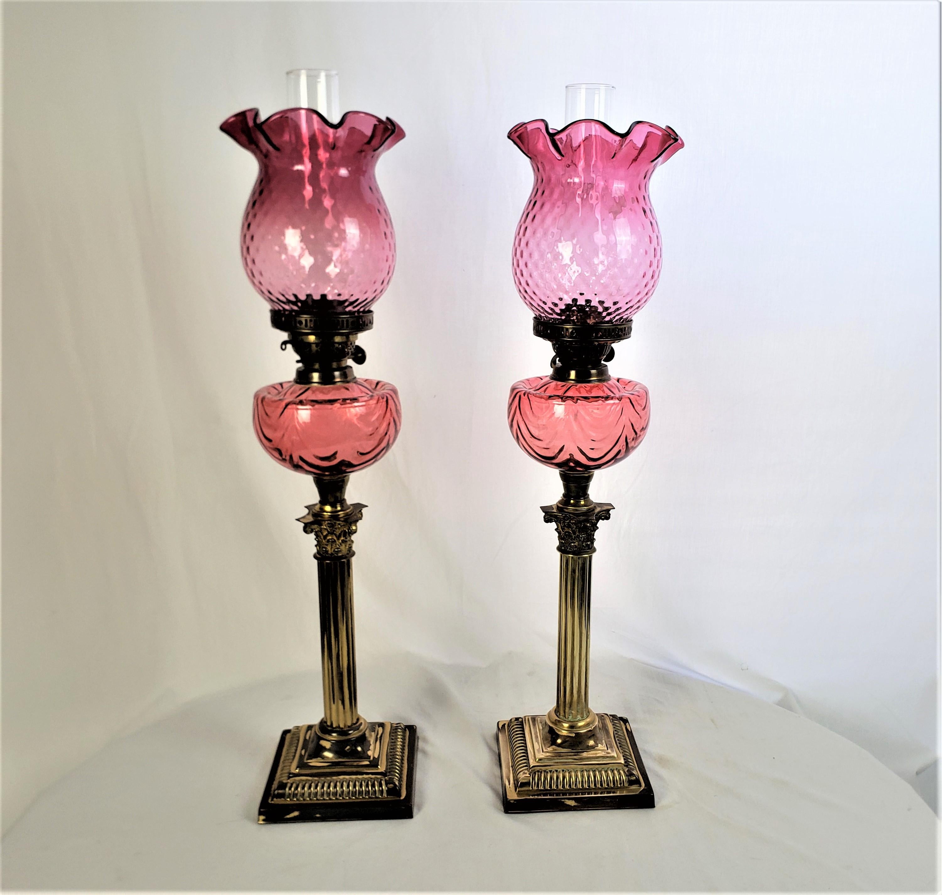 This pair of antique banquet or parlor lamps likely originate from England and date to approximately 1880 and done in the period Victorian style. The lamp bases are done in brass and are done with a Corinthian column style. The outer shade is done