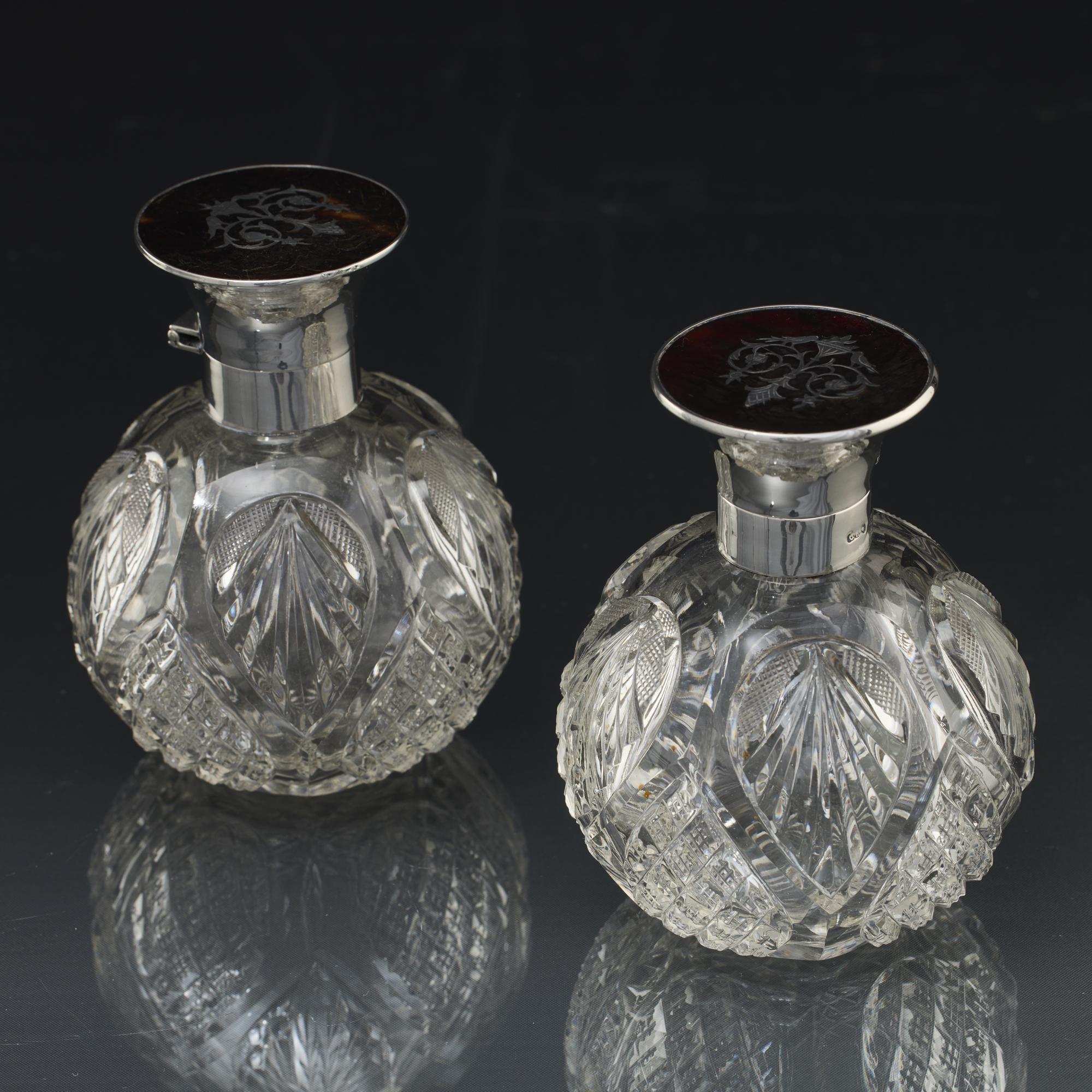 A pair of early-20th-century antique perfume or scent bottles with hand-cut glass bodies decorated with lattice and fanned flute panels, as well as sections of diamond pattern. There is a starburst cut into each base. The bottles have silver