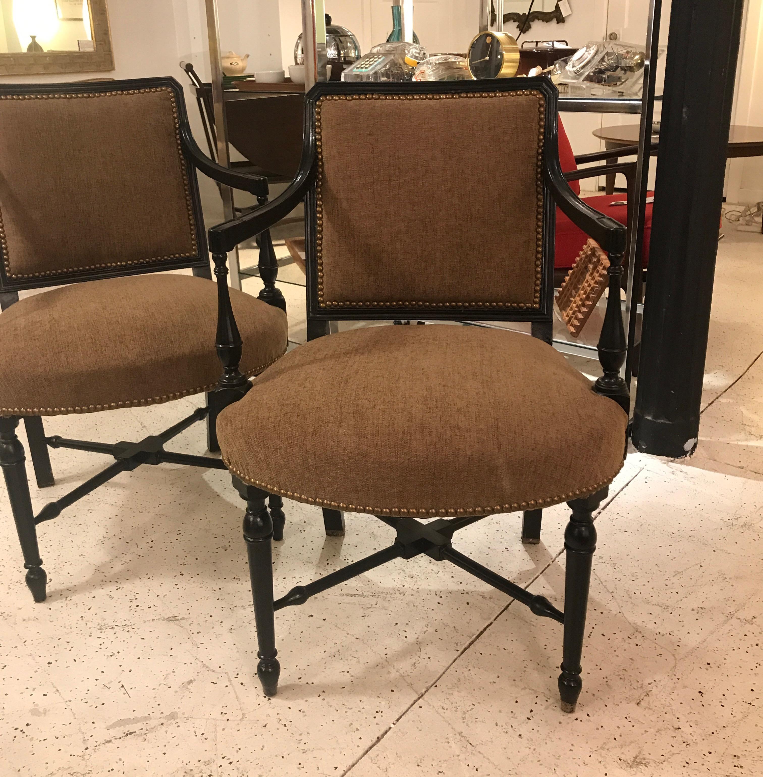 A pair of ebonized armchairs with neutral olive-tan chenille upholstery. The sturdy wood frame with ebony finish is trimmed with antiqued brass nail head trim. Handsome shape with nicely turned legs and X-stretcher base and slightly sloping arms.