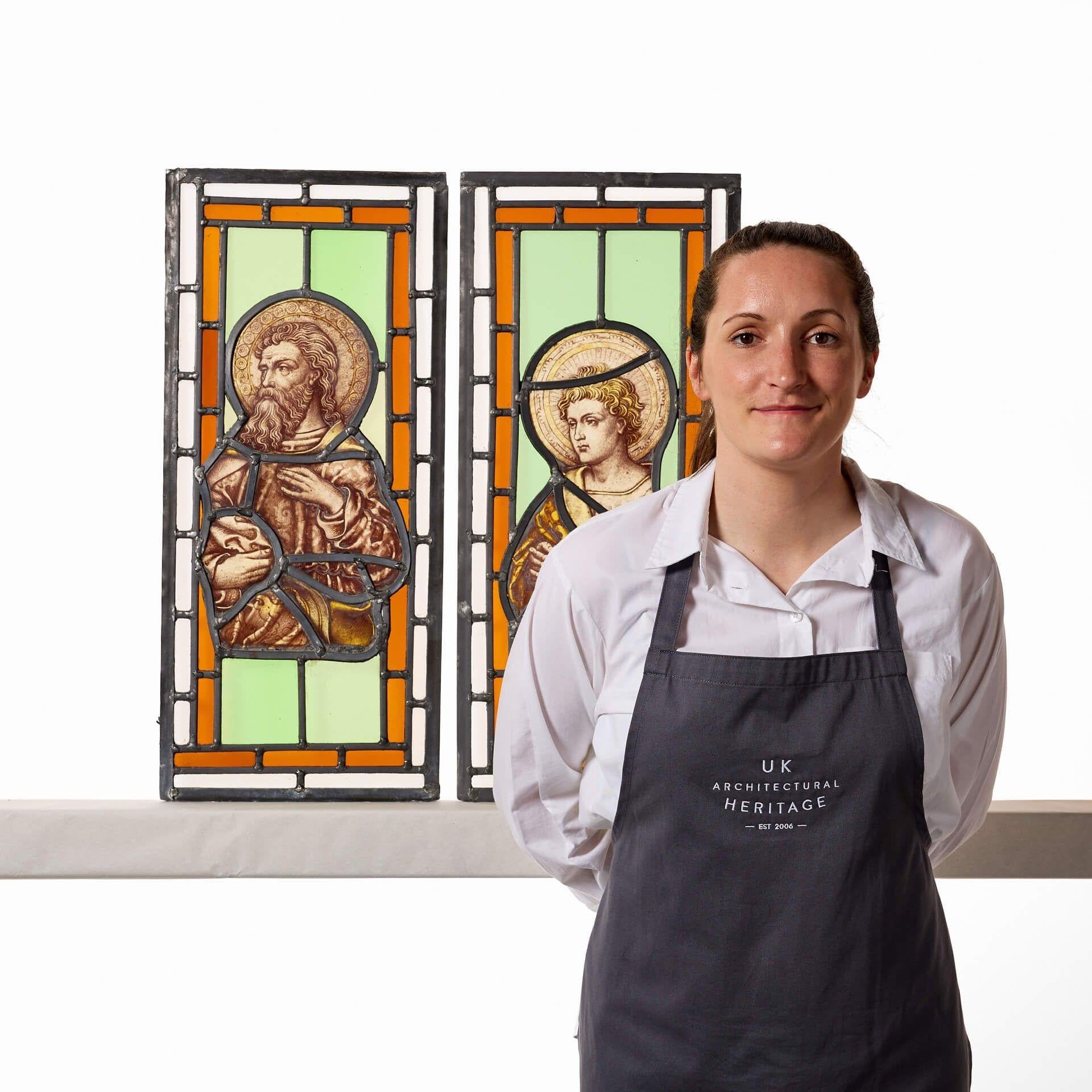 A pair of antique ecclesiastical stained glass windows circa 1850 depicting two saintly figures: one a bearded, long-haired man and the other a young boy with locks of blonde hair. Each is set into a backdrop of vibrant green and orange glass,