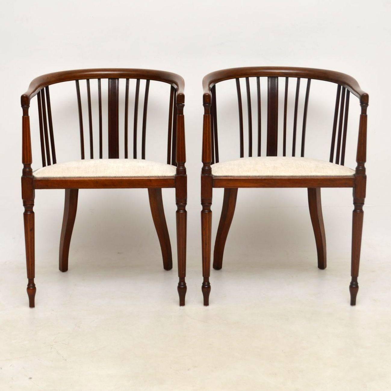 Elegant pair of antique Edwardian mahogany tub back armchairs in good condition and upholstered in a neutral color fabric. The frames are finely inlaid with satinwood on the backs, arms and legs. They have curved backs and sit on turned legs. These
