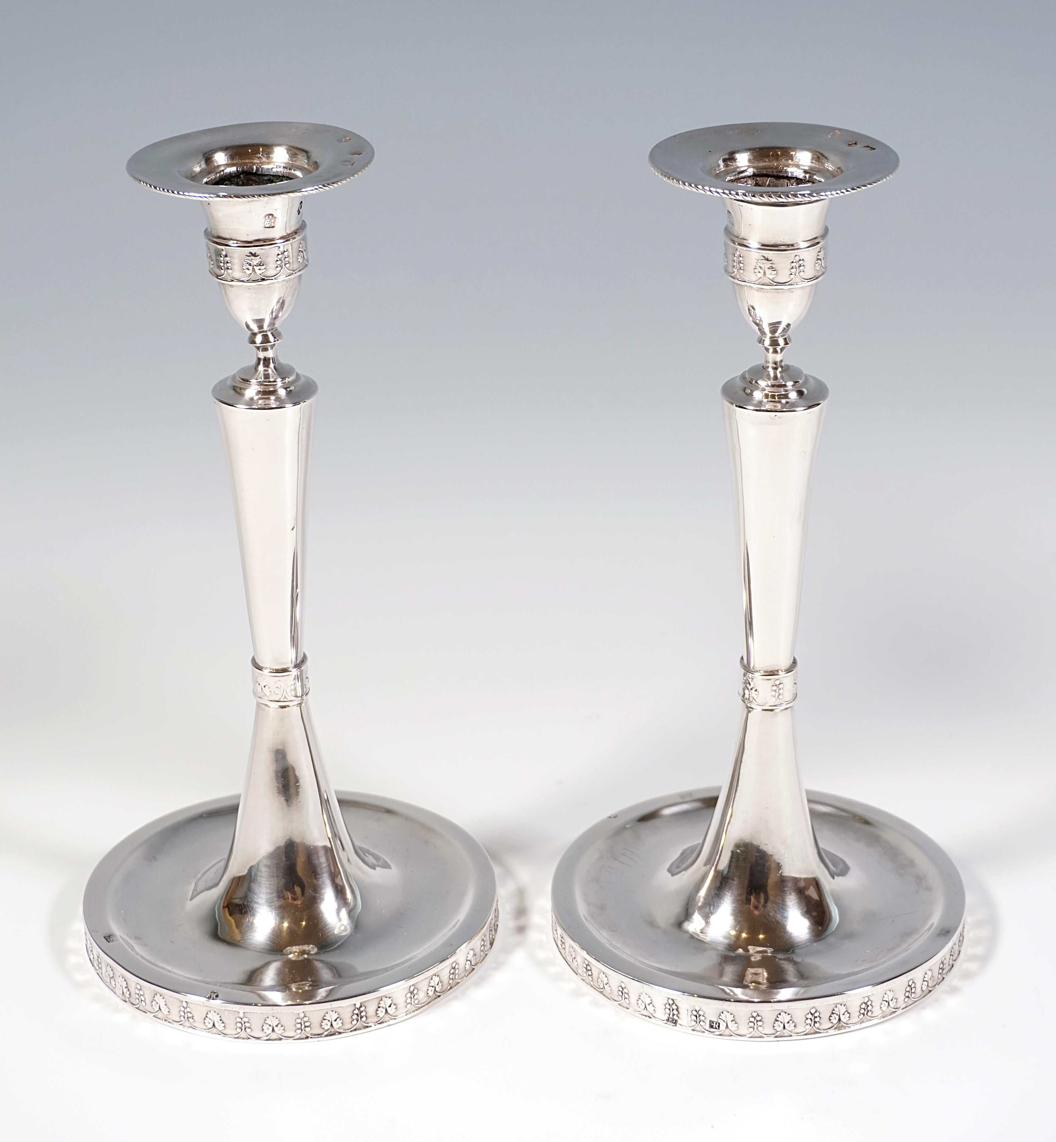 Two elegant, simple silver candlesticks on a round base, the smooth shaft raised in the centre with a constriction formed by a beaded band in relief, the shaft widening conically towards the top with a vase-shaped spout on top, relief bands on the