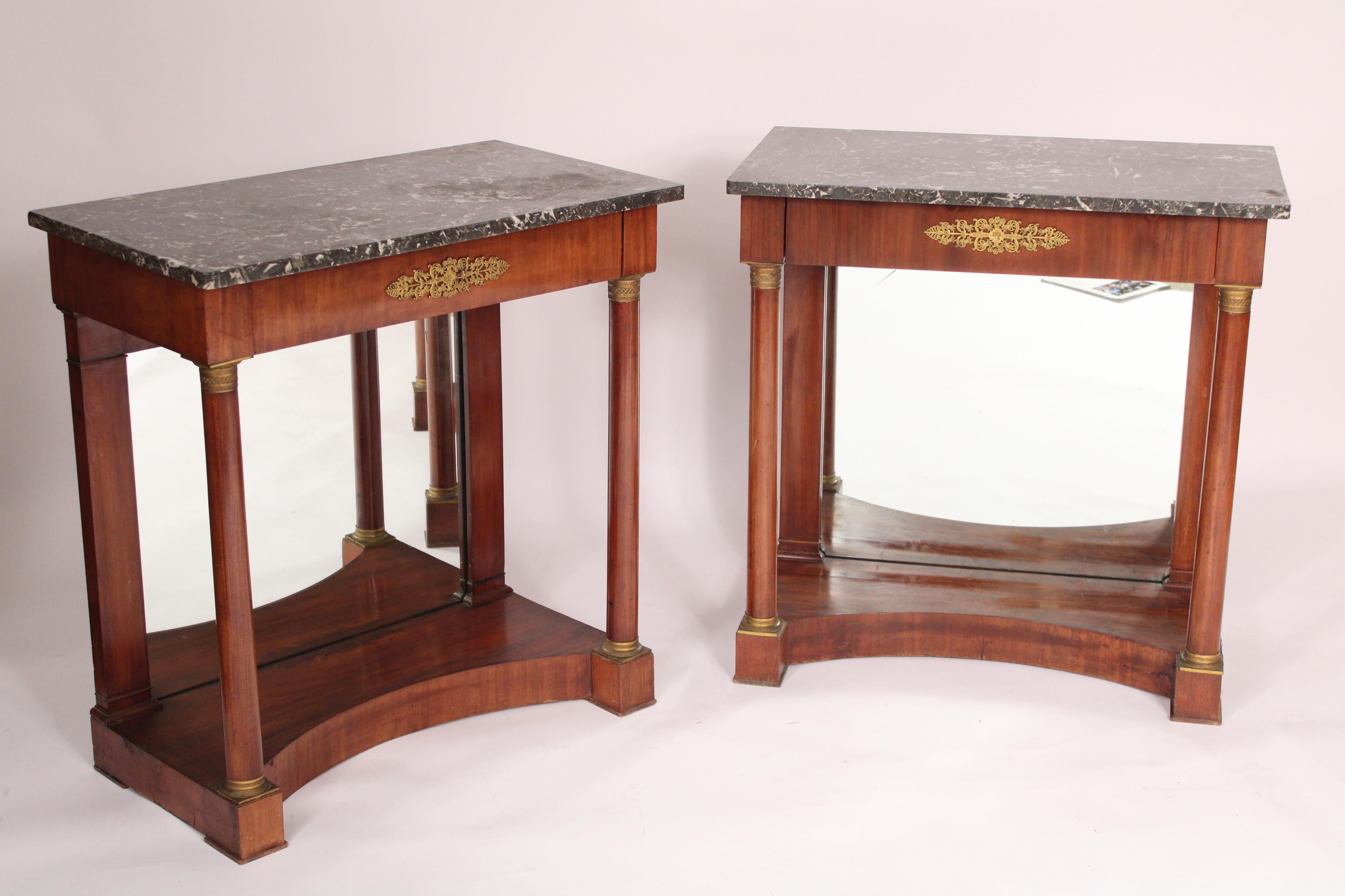 Pair of antique Empire style brass mounted mahogany console tables with marble tops, circa 1900. With grey marble tops, frieze drawer with a central classical brass mounting, two front columns with brass mountings on capitals and plinths and a