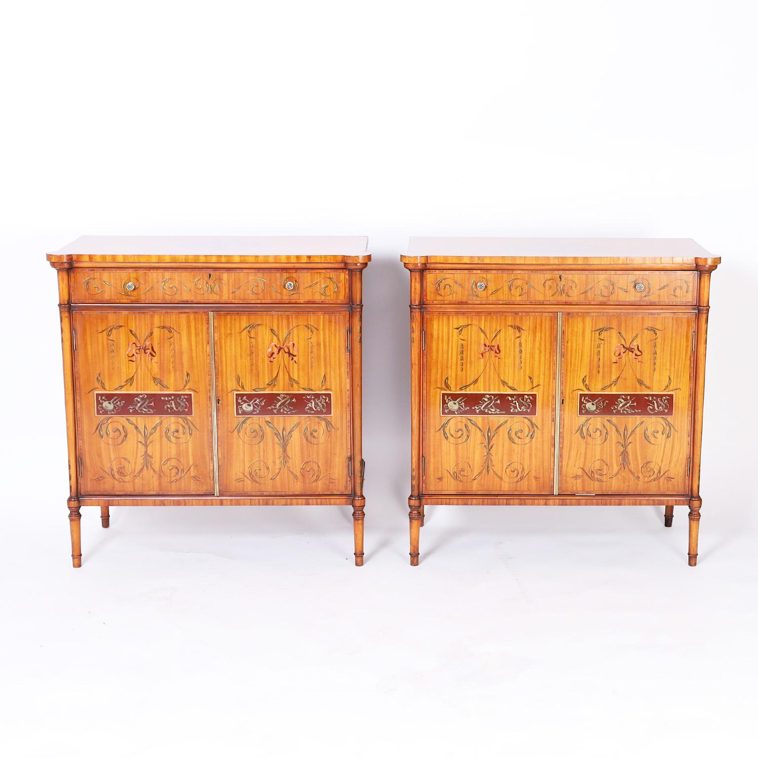 Adams style cabinets crafted in satinwood featuring hand painted musical medallions on the tops, a drawer and two doors decorated with instruments, leaves and ribbons, hand dovetailed construction and elegant turned legs.
