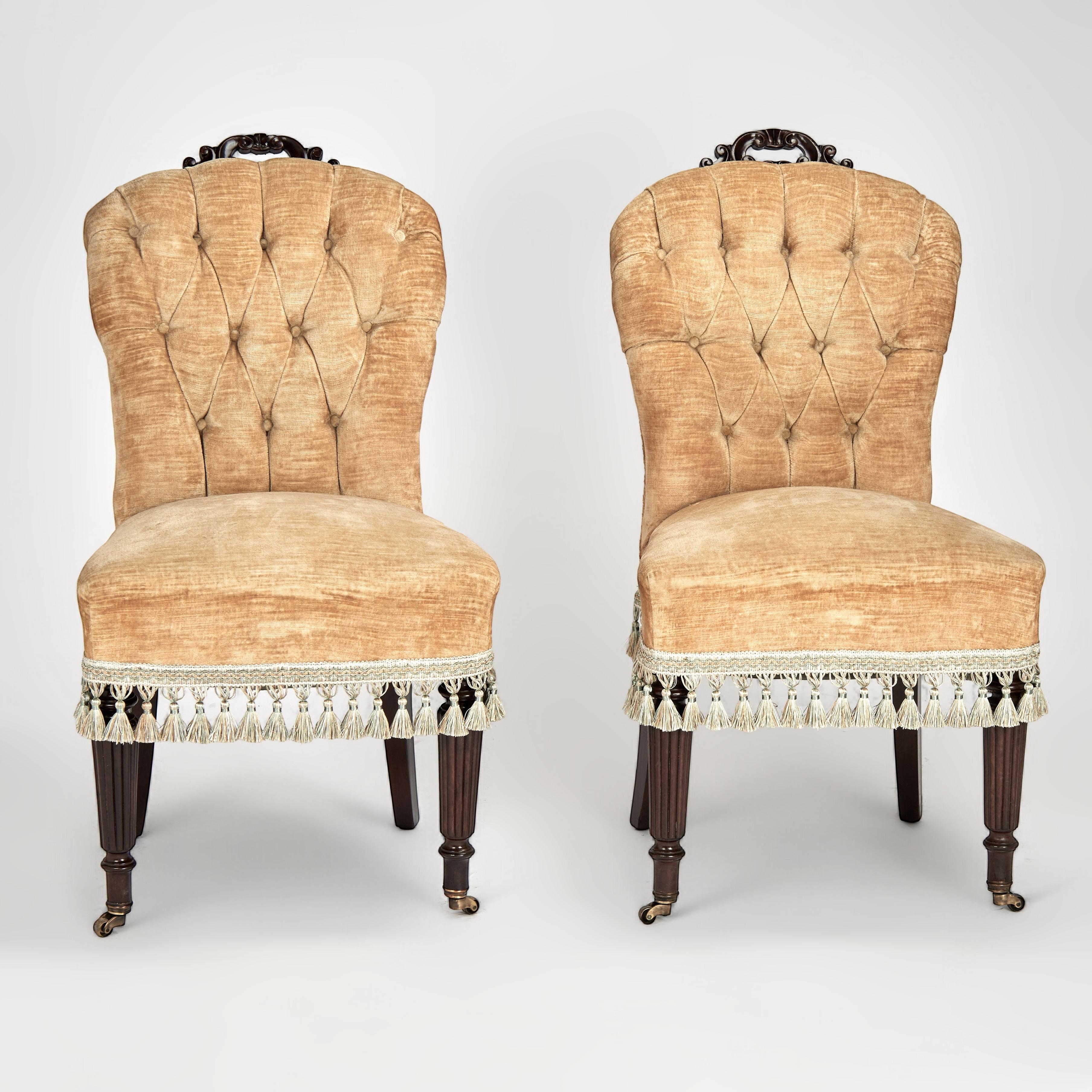 Affascinante coppia di poltrone da salotto o da boudoir inglesi del XIX secolo, realizzate in mogano e di forma minuta, con maniglie floreali intagliate sui piani e imbottitura in velluto con schienali a bottoni e sedili con nappe su gambe scanalate
