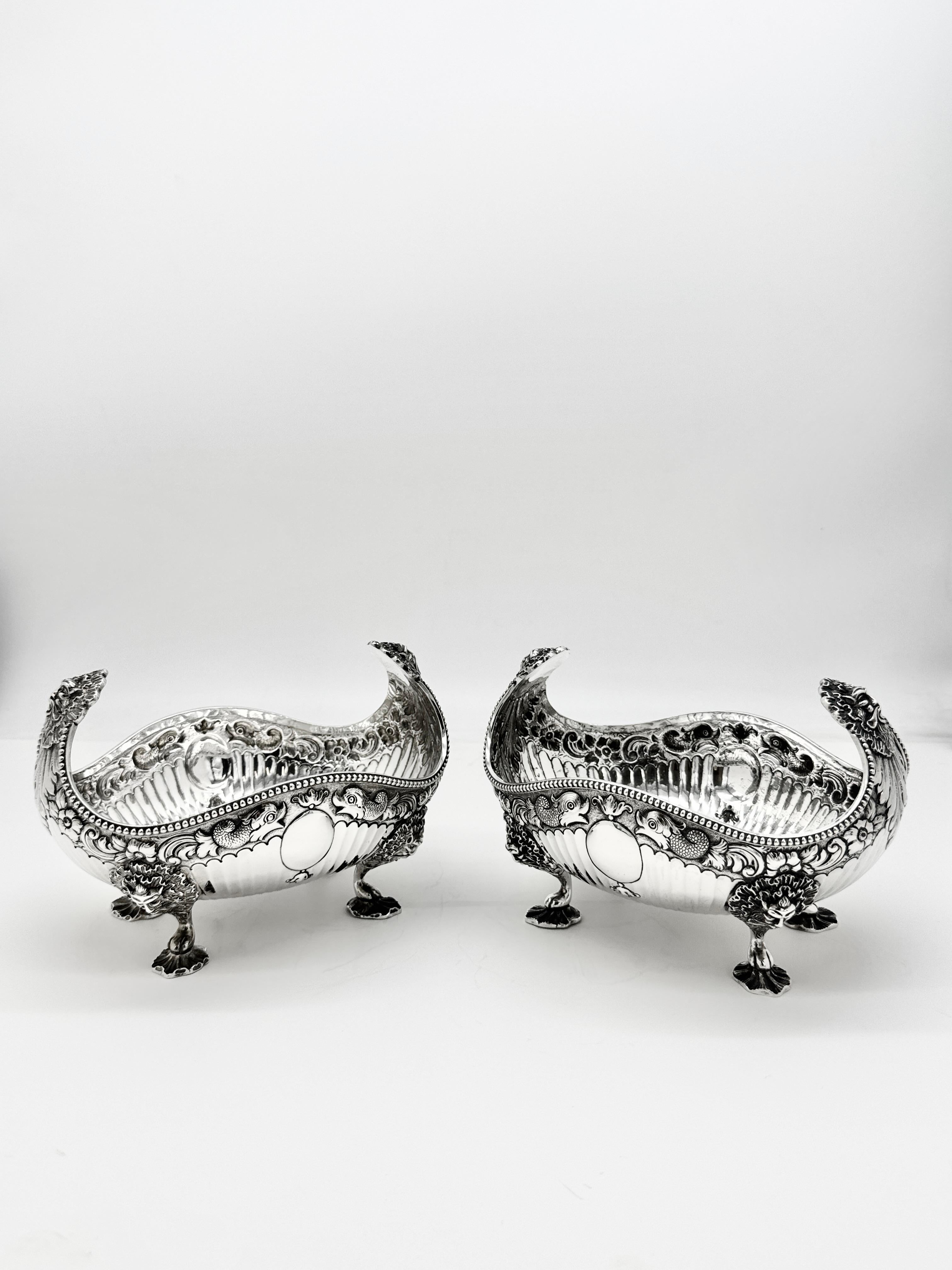 Paire de plats à fruits en argent sterling de forme ovale, ajourés et estampés de feuillages, de motifs de poissons et de cartouches vacants, reposant sur quatre pieds en forme de masque de lion et de patte d'oie. Les poignées se terminent par des