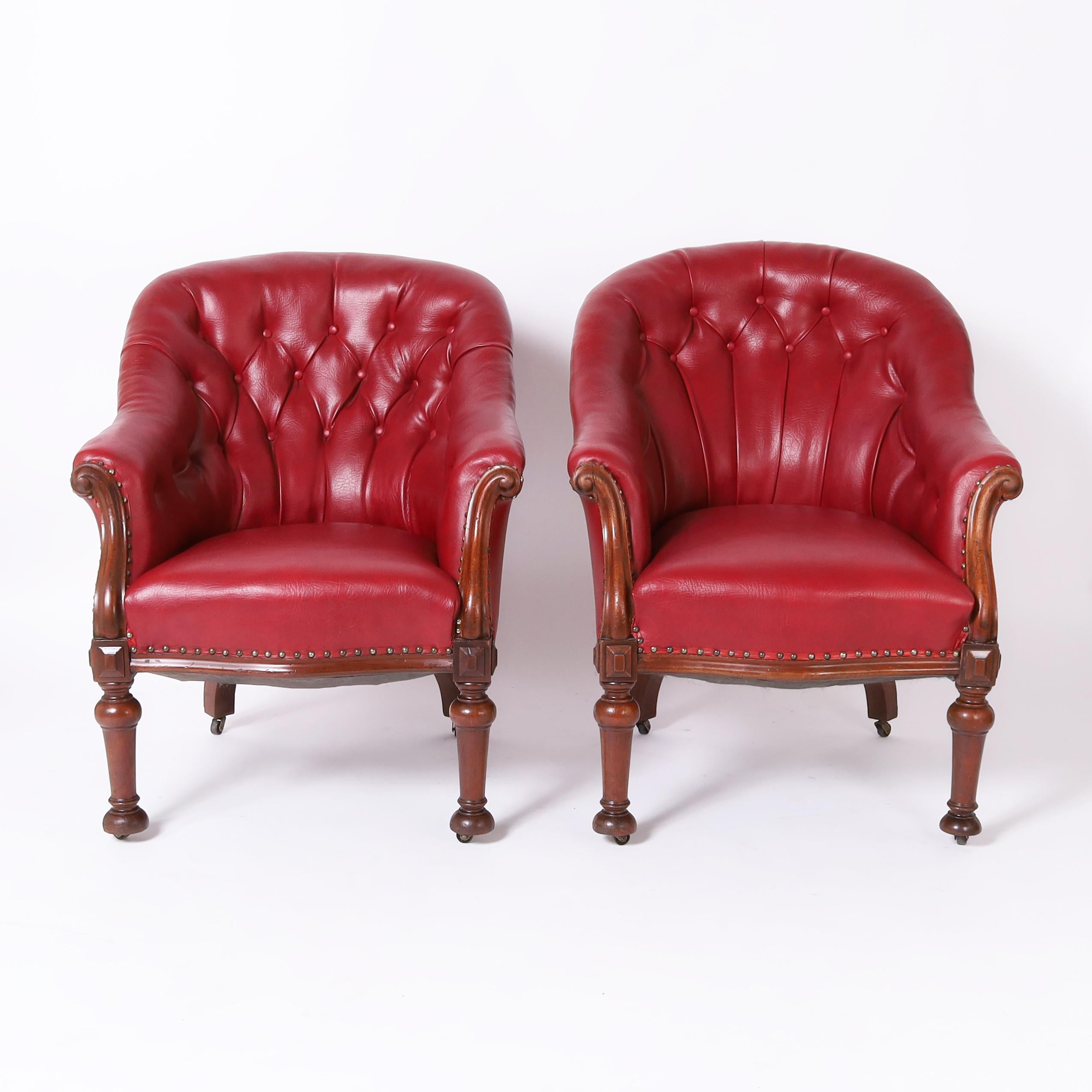 Handsome pair of lady and gentleman William IV English club chairs handcrafted in mahogany featuring alluring red faux leather button tufted upholstery, elegant arms, and turned front legs on casters.
