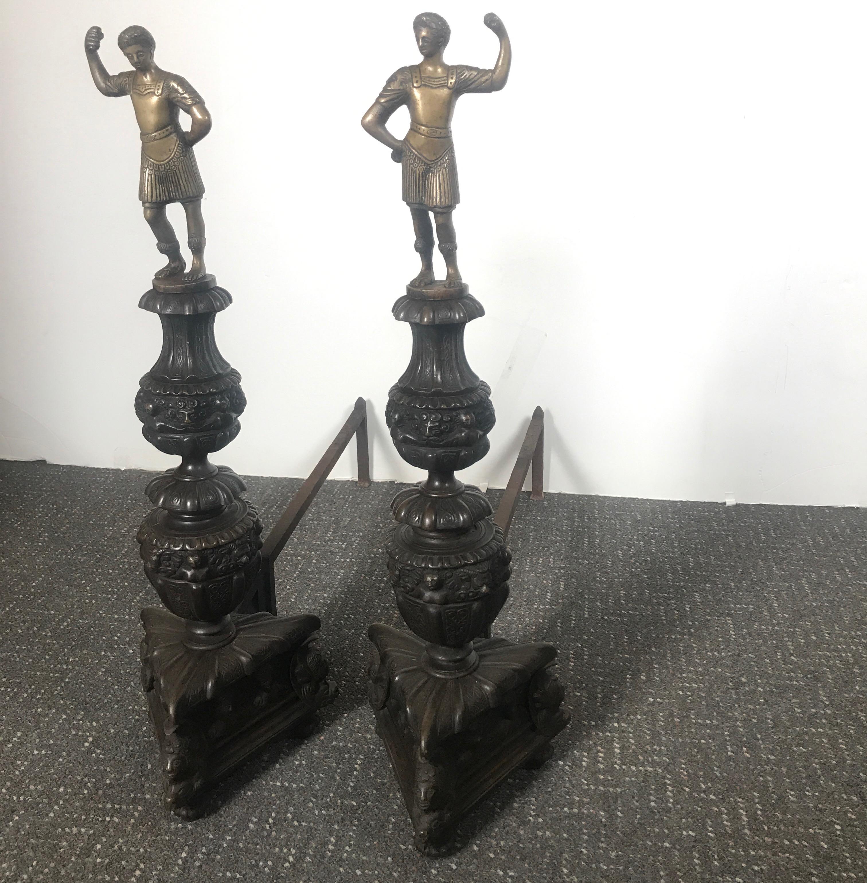Antique bronze andirons with wrought iron backs. The tall figurative andirons with a Roman figure on the top above finely cast elaborate bases with triangular bases.