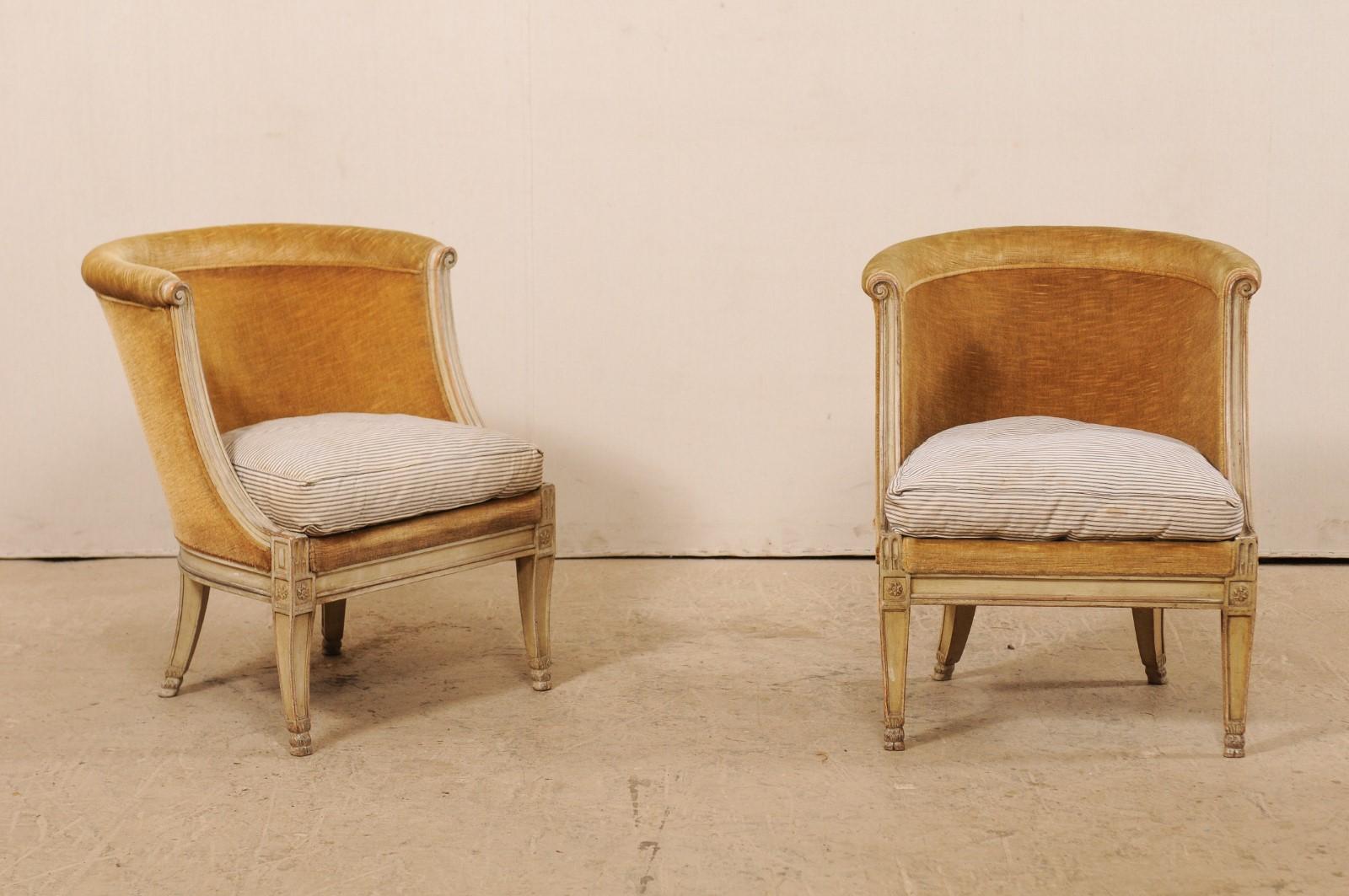 A pair of French barrel back club chairs from the early 20th century. This antique pair of bergères occasional chairs from France have upholstered and barrel backs with rounded top rail, and volute carvings at either top side. The seat rail is