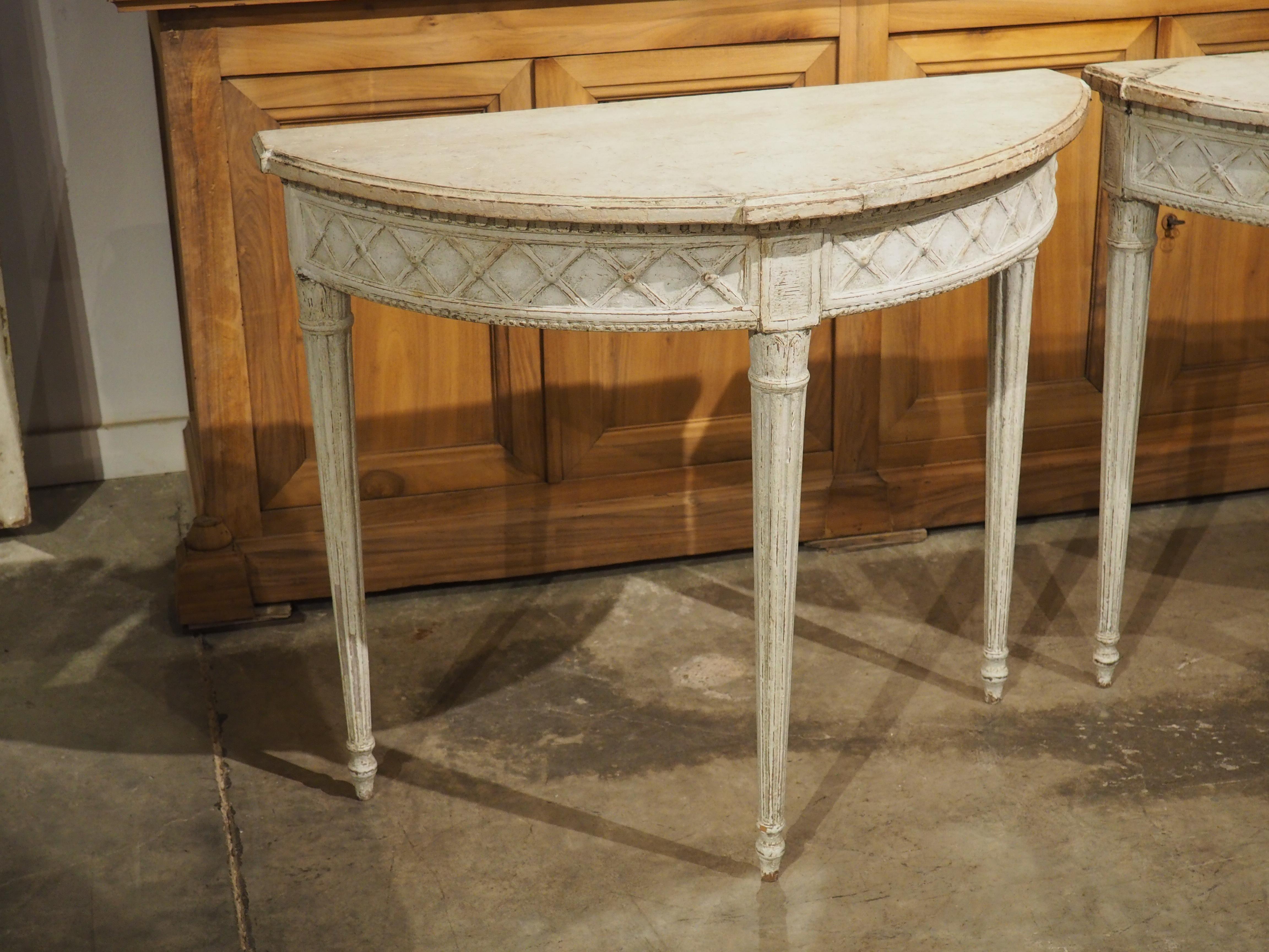 This pair of painted demi-lune tables were hand-carved, circa 1880s, in France in the style of Louis XVI. The statuesque tables feature a more recent white finish with areas that have developed a gray patination.

True to the style that prevailed