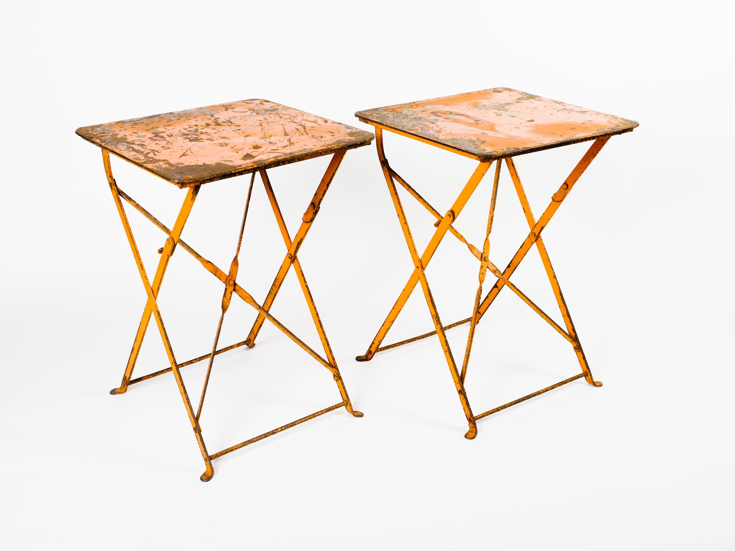 Pair of industrial French folding iron bistro tables with beautiful patina on the metal and original orange paint. Exhibits wear on the paint throughout, adding to their rustic charm and beauty. Can be used indoors as side tables or outdoors as