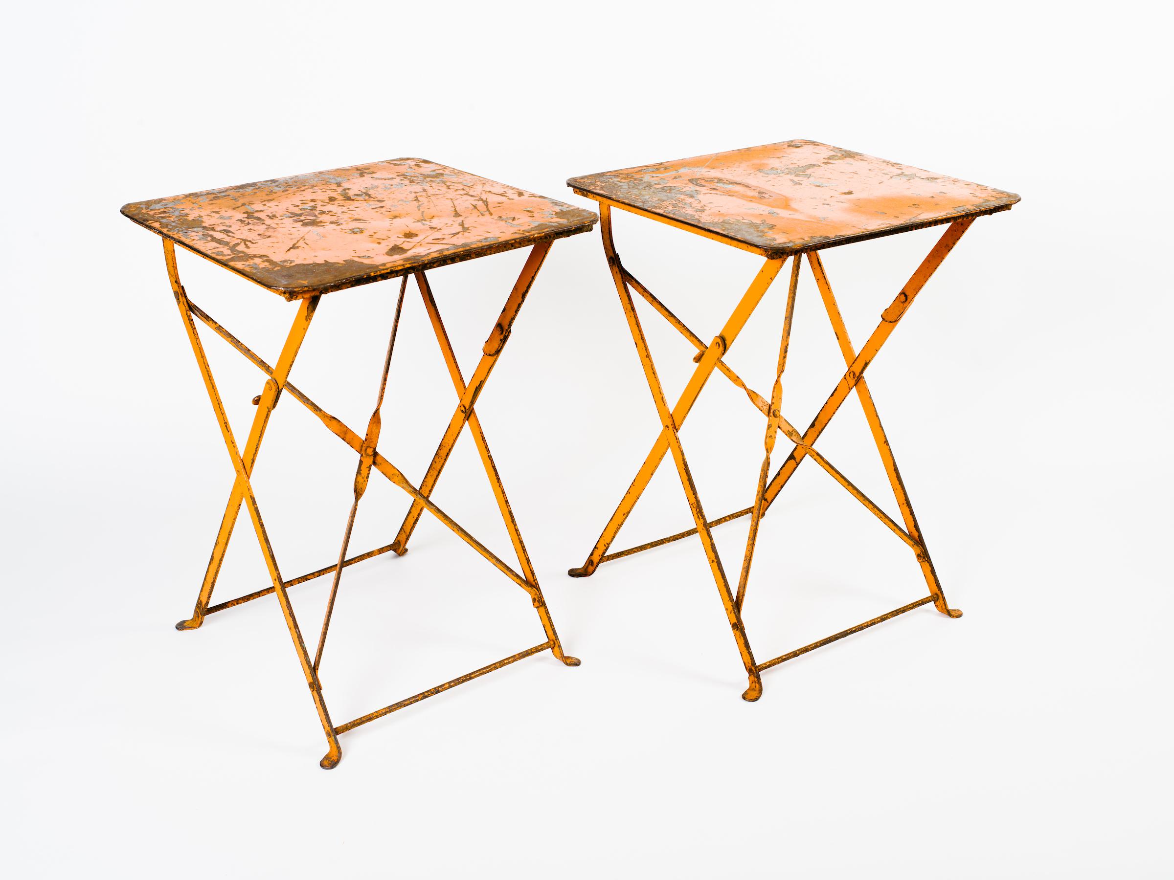 Pair of 1930s industrial French folding iron bistro tables with beautiful patina on the metal and original orange paint. Exhibits wear on the paint throughout, adding to their rustic charm and beauty. Can be used indoors as side tables or outdoors