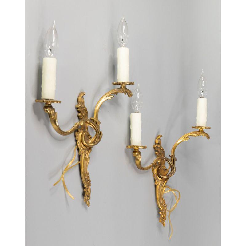 A gorgeous pair of antique French gilded bronze candelabra two arm light wall sconces. These beautiful sconces have a lovely patina with gilt scrolls, acanthus leaves, and wax candle sleeves. They are are solid and heavy and the wiring is in