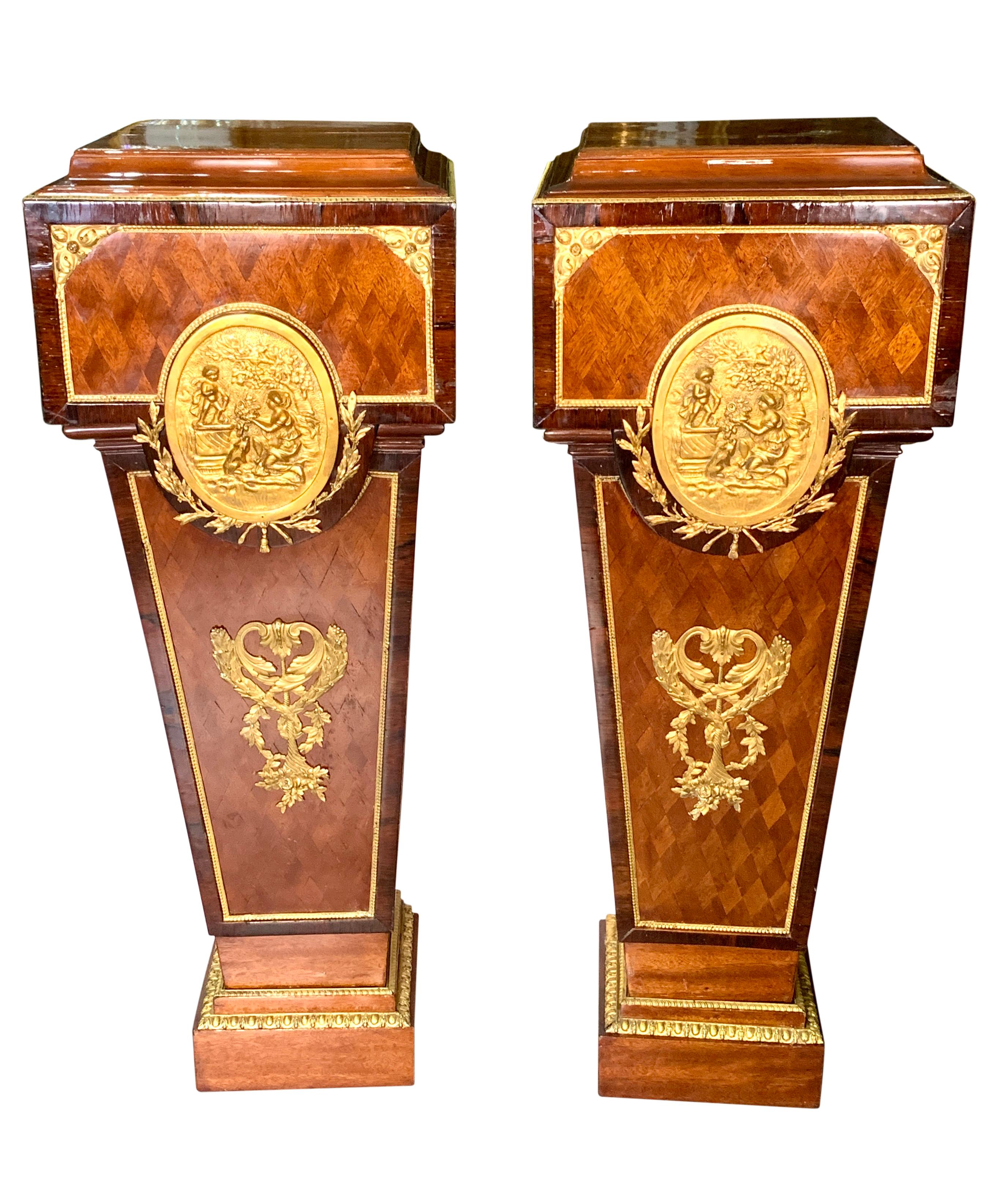 An elegant pair of early 20th century French Louis XVI style gilt bronze mounted pedestals. Constructed in parquetry wood with gilt bronze mounts, rising from stepped rectangular plinths, with a central oval gilt bronze plaque depicting a lady