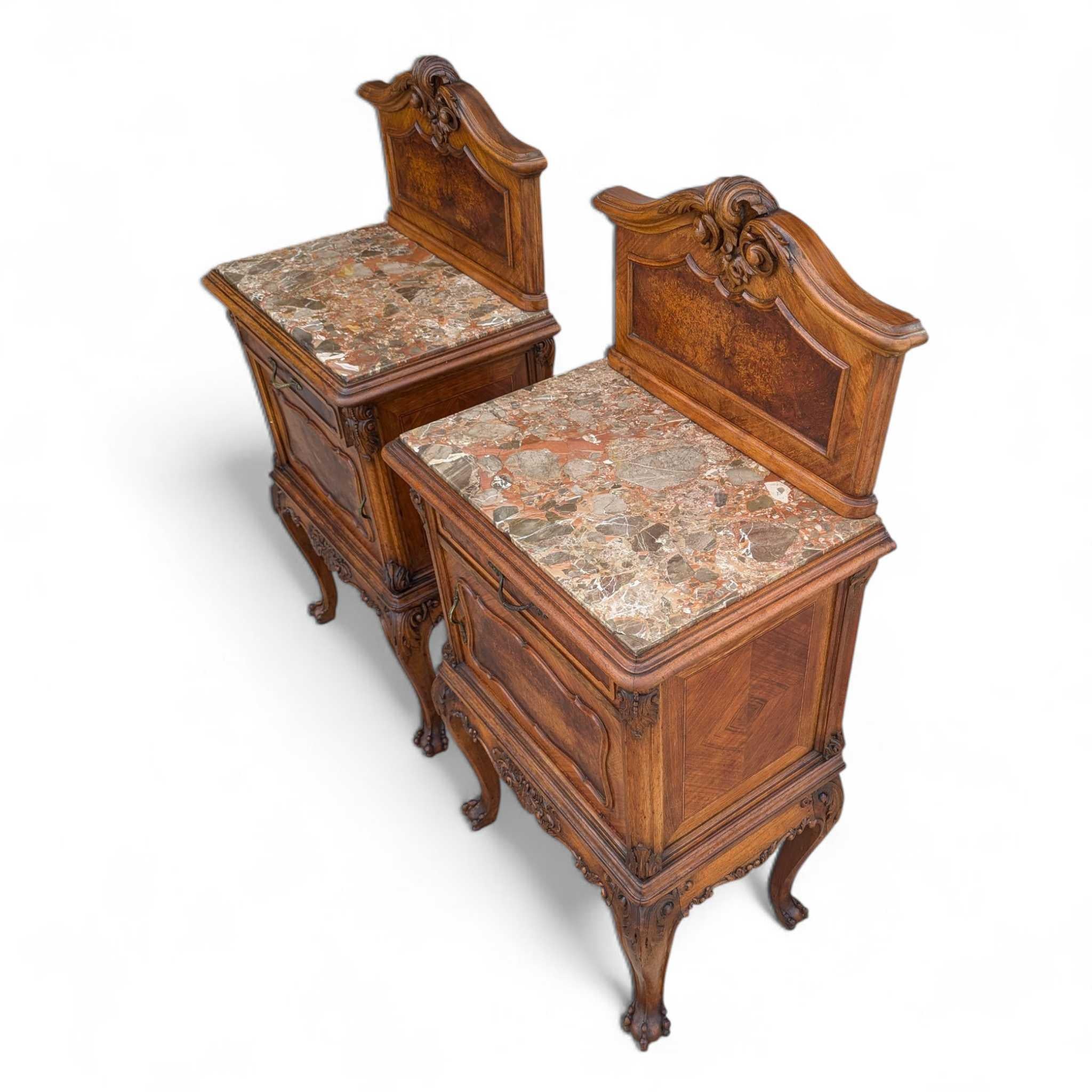 A pair of antique French Louis XV style nightstands with variegated marble tops, circa 1900. Each stand features a carved crest with foliate scrolls, burled wood panels framed in walnut, and floral carvings on the apron and cabriole legs. One drawer