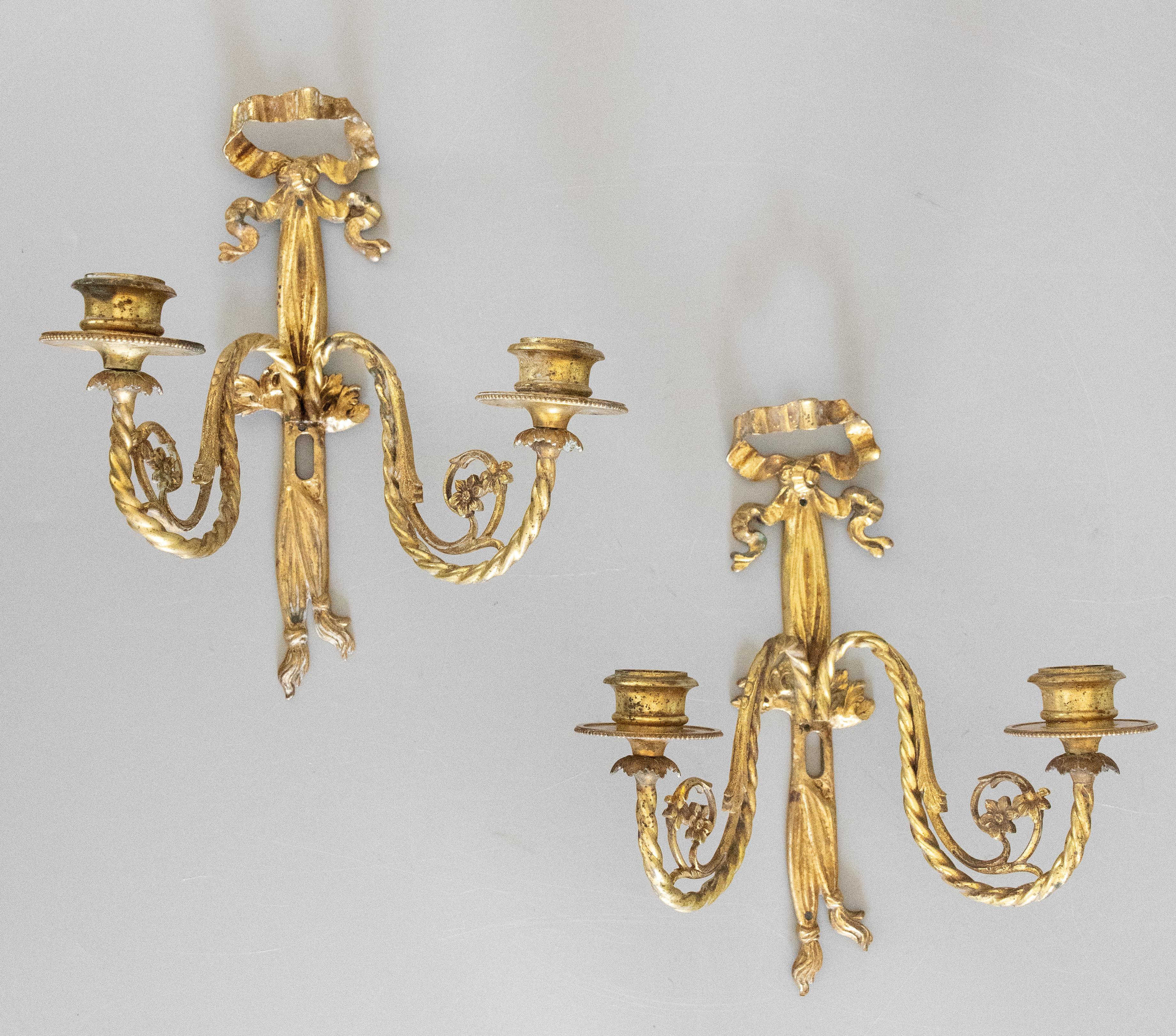 A gorgeous pair of antique early 20th-Century French Louis XVI style gilded bronze two arm candle wall sconces, sourced in Paris. These beautiful sconces are decorated with gilt twisted arms, flowers, bows and ribbons with tassels in a lovely gilt