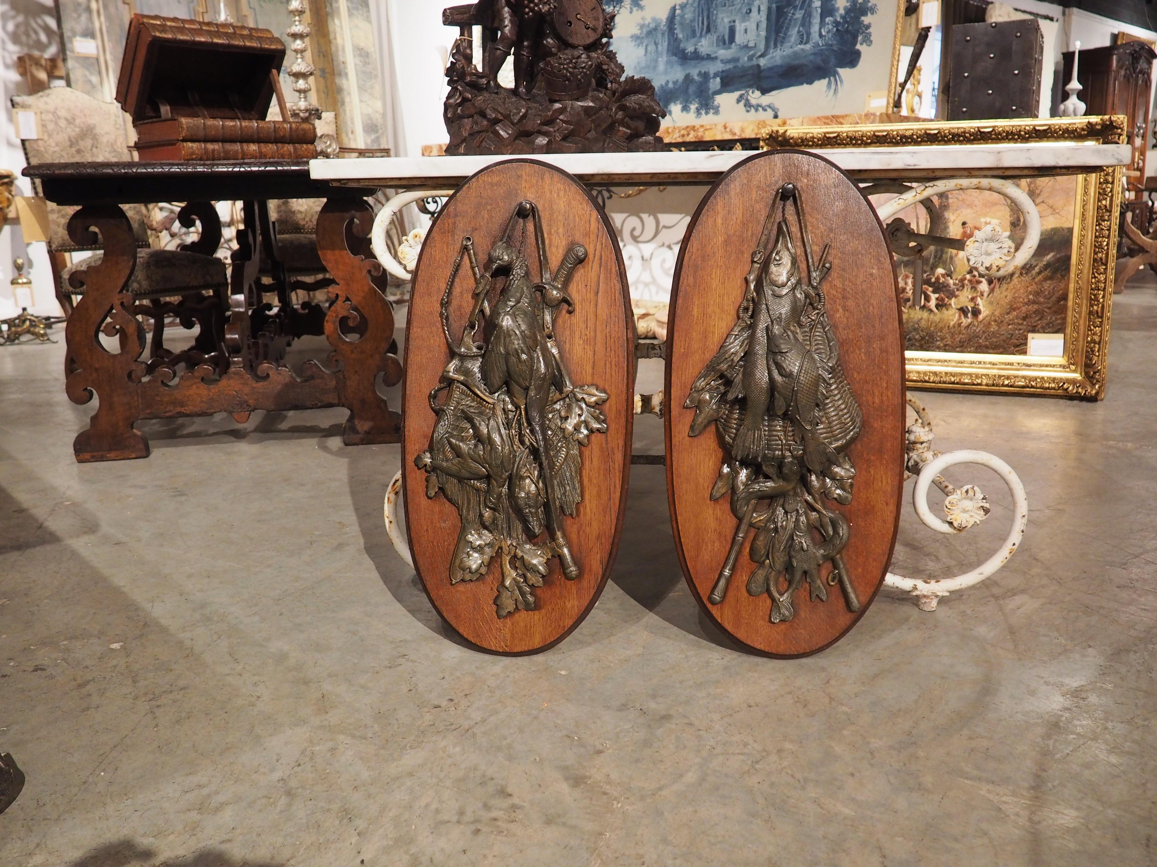 From France, circa 1900, this pair of regule trophy plaques have been mounted on oval, carved oak backings, which has a thin cavetto molding. One of the trophies is hunting-inspired, with a trio of felled birds in front of a tasseled hunting bag,
