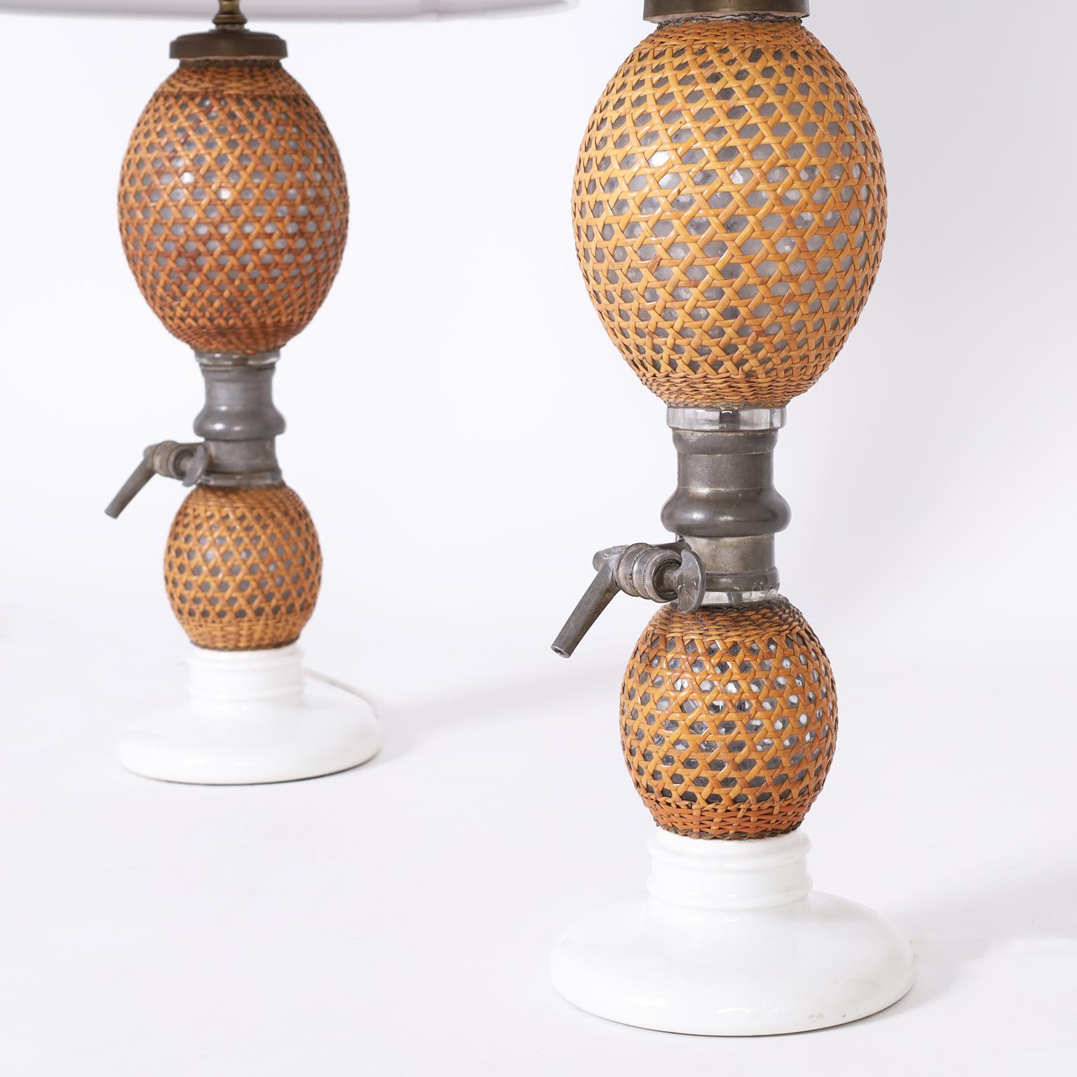 Antique French seltzer bottles now table lamps with double glass bulbs wrapped with wicker, pewter spout and valve mechanism, and glazed white porcelain bases.