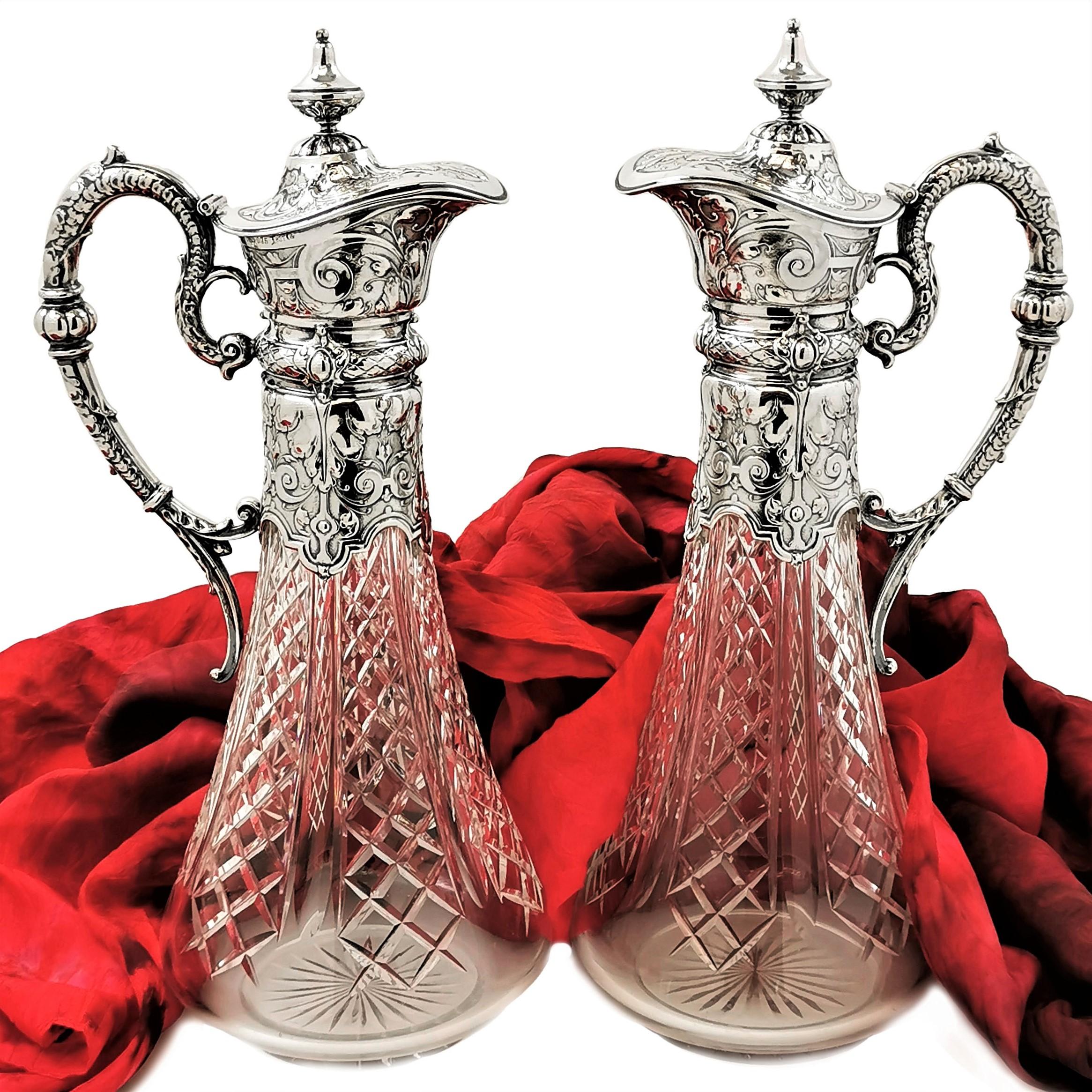 An elegant pair of Antique German Solid Silver & Glass Claret Jugs with solid silver mounted necks, lids and handles on cut glass bodies. The silver necks and lids of the Jugs are decorated with ornate chased patterns and the design on the handle