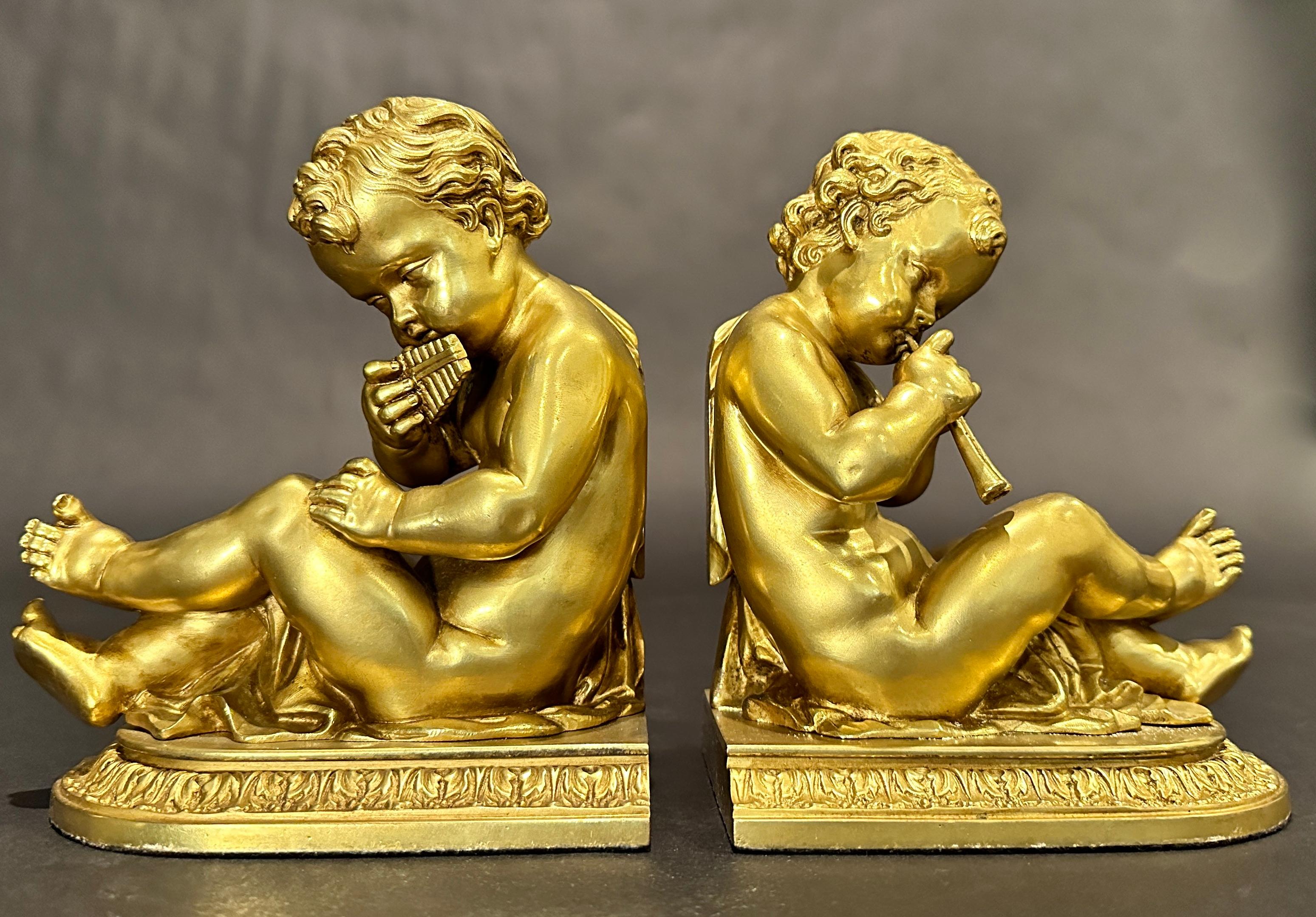Paire de serre-livres anciens en bronze doré. Chacune représente un jeune enfant/putti jouant d'un instrument de musique. L'un est un syrinx (d'après l'instrument conçu par le dieu Pan), l'autre un Aulos (flûte à anche double), assis sur une