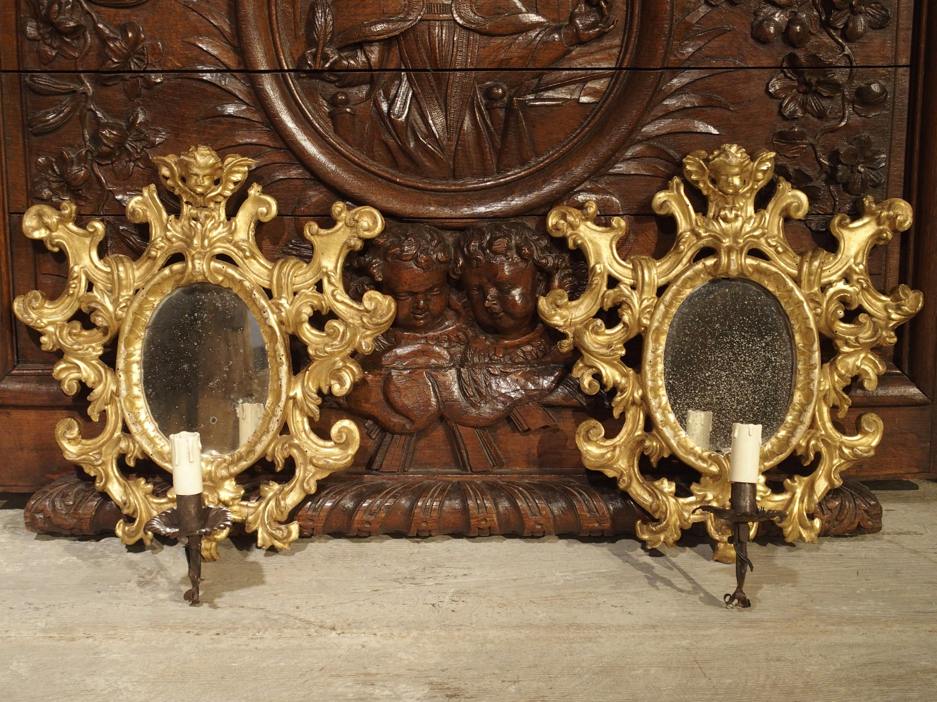These lovely antique Italian single arm giltwood sconces date to the late 1800s. They have oval mirrors with lovely cherub motifs at the top center. The motifs surrounding the mirrors are curled C-and S-Scrolls with acanthus leaf motifs curling