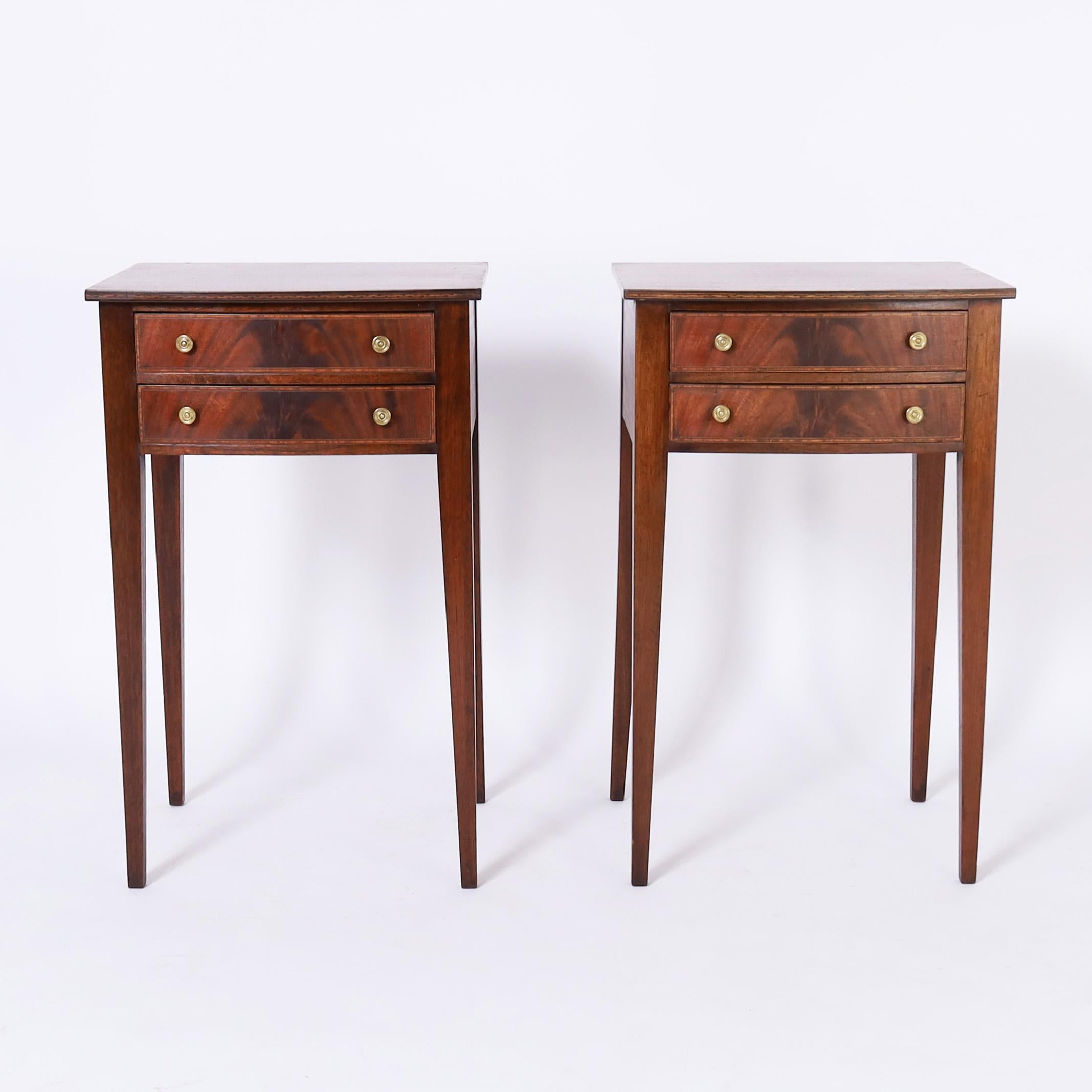 Standout pair of early 19th century English stands handcrafted in mahogany with bow front tops having geometric inlaid edges over two drawer cases with flame mahogany fronts and cross banded inlays on elegant tapered legs.