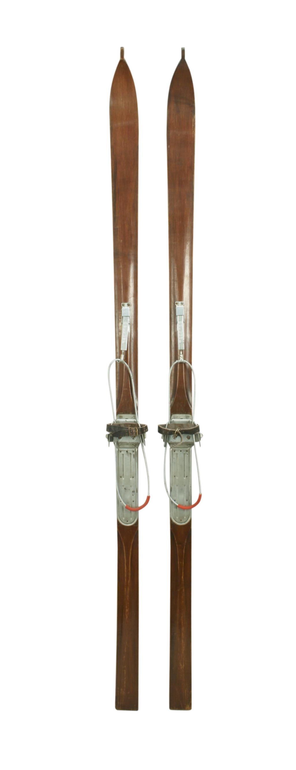 Ash ridge top skis with galvanised toe irons and foot plates, leather foot straps and and cable bindings. The skis are with pointed tips, dark brown in colour with a polished finish and the running surface having a single groove along its length.