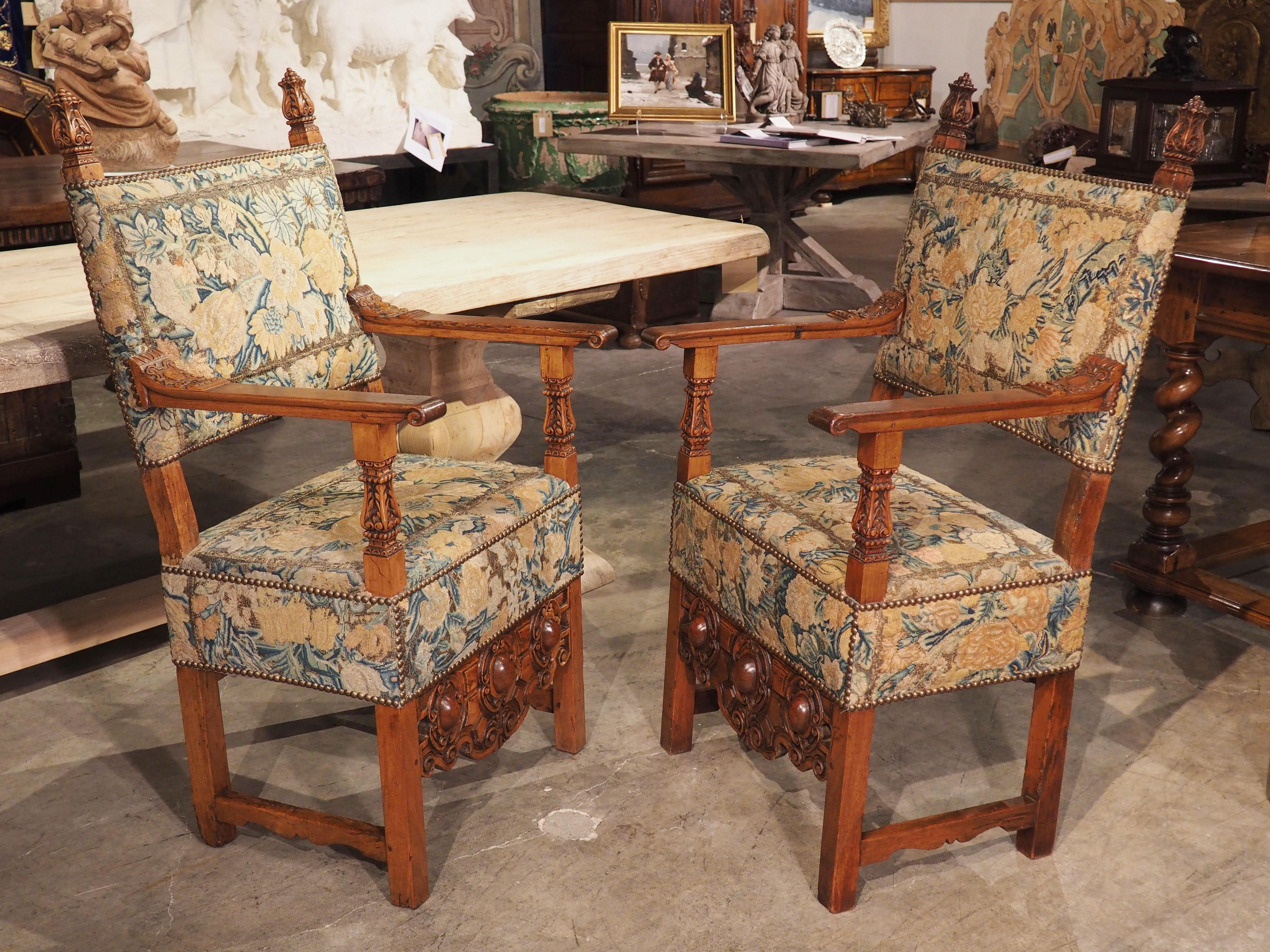 Hand-carved in Italy in the 1600’s, this pair of elegant walnut armchairs features a rich foliate and floral needlepoint upholstery. The chairbacks, seats and aprons of each chair has been covered by embroidery patterned with green, gold, and pink