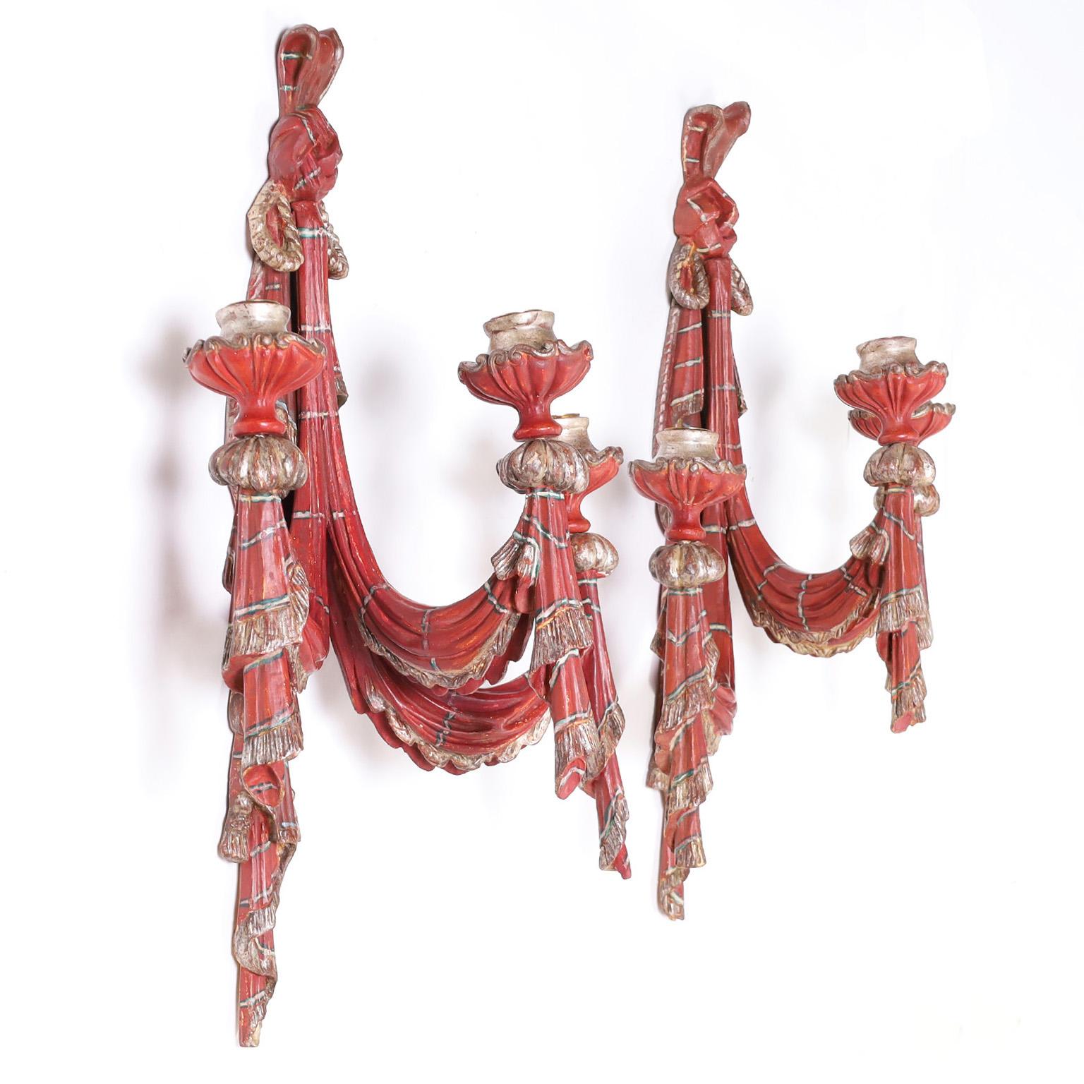 Antique pair of Venetian style three light Italian wall sconces carved from hardwood in an elegant faux drapery composition decorated with an alluring red. Highlighted with silver leaf rope and tassel, all worn to perfection.