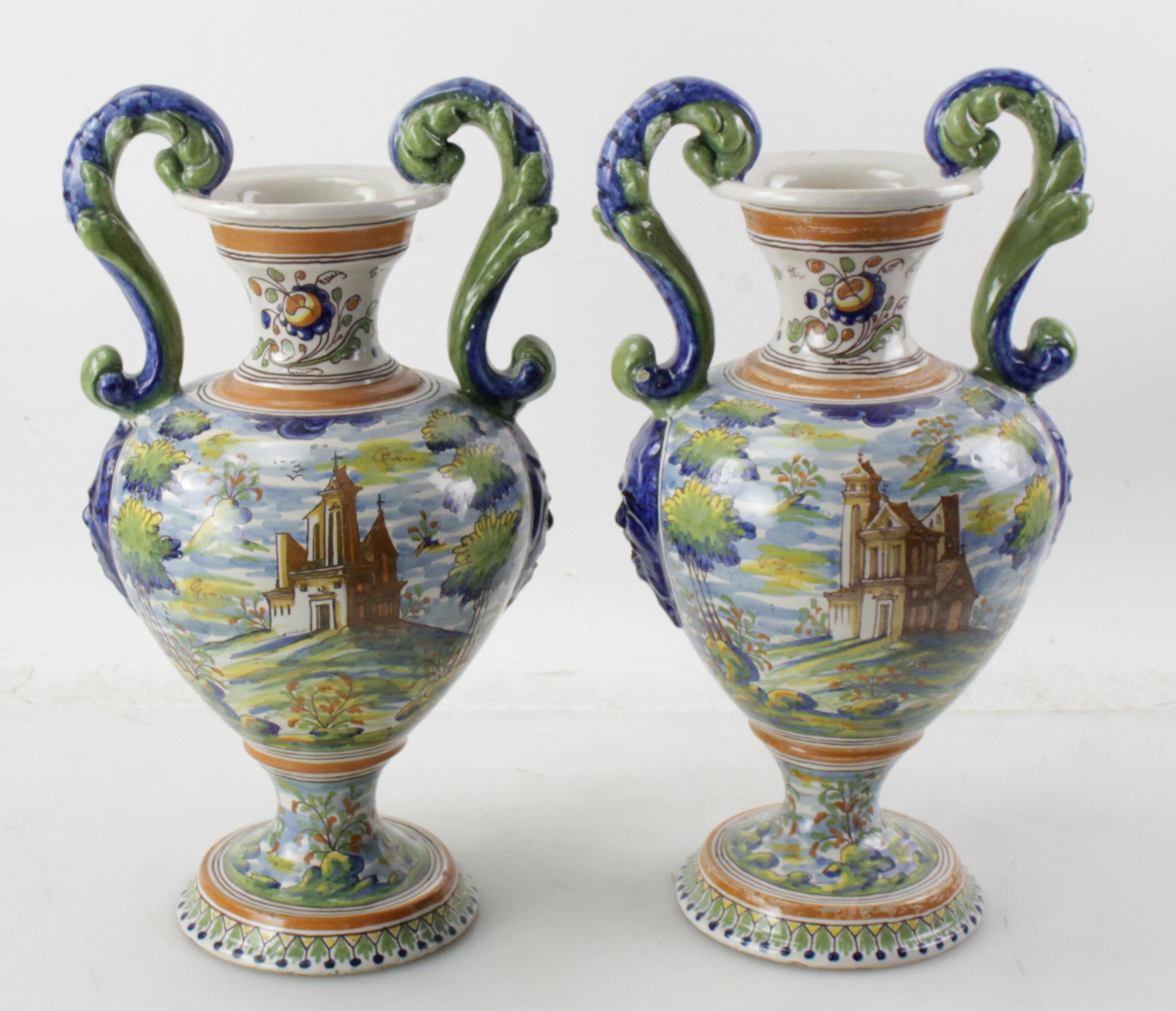 
From Italy, this colorful pair of antique Majolica urns have different, hand-painted,  scenes on each urn.  On the bodies are brightly colored scenes with churches, figures, and landscaping from Grecian or Roman times. A lovely pair of antique