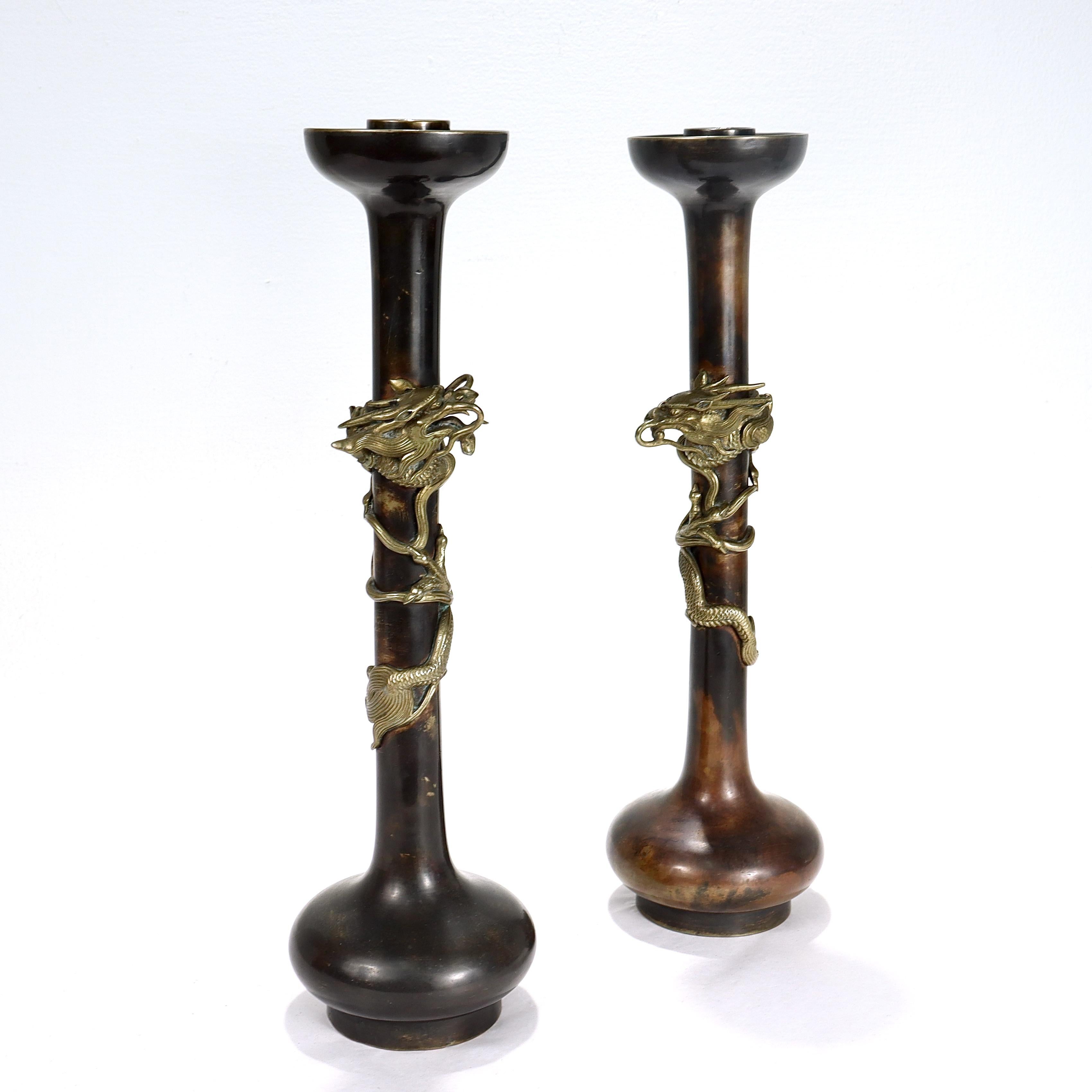 A fine pair of antique Japanese bronze candlesticks.

Each decorated with an applied, coiled dragon to its elongated thin neck below an everted lip and candle cup and above a suppressed ball foot and base. Each dragon with highly detailed hammered