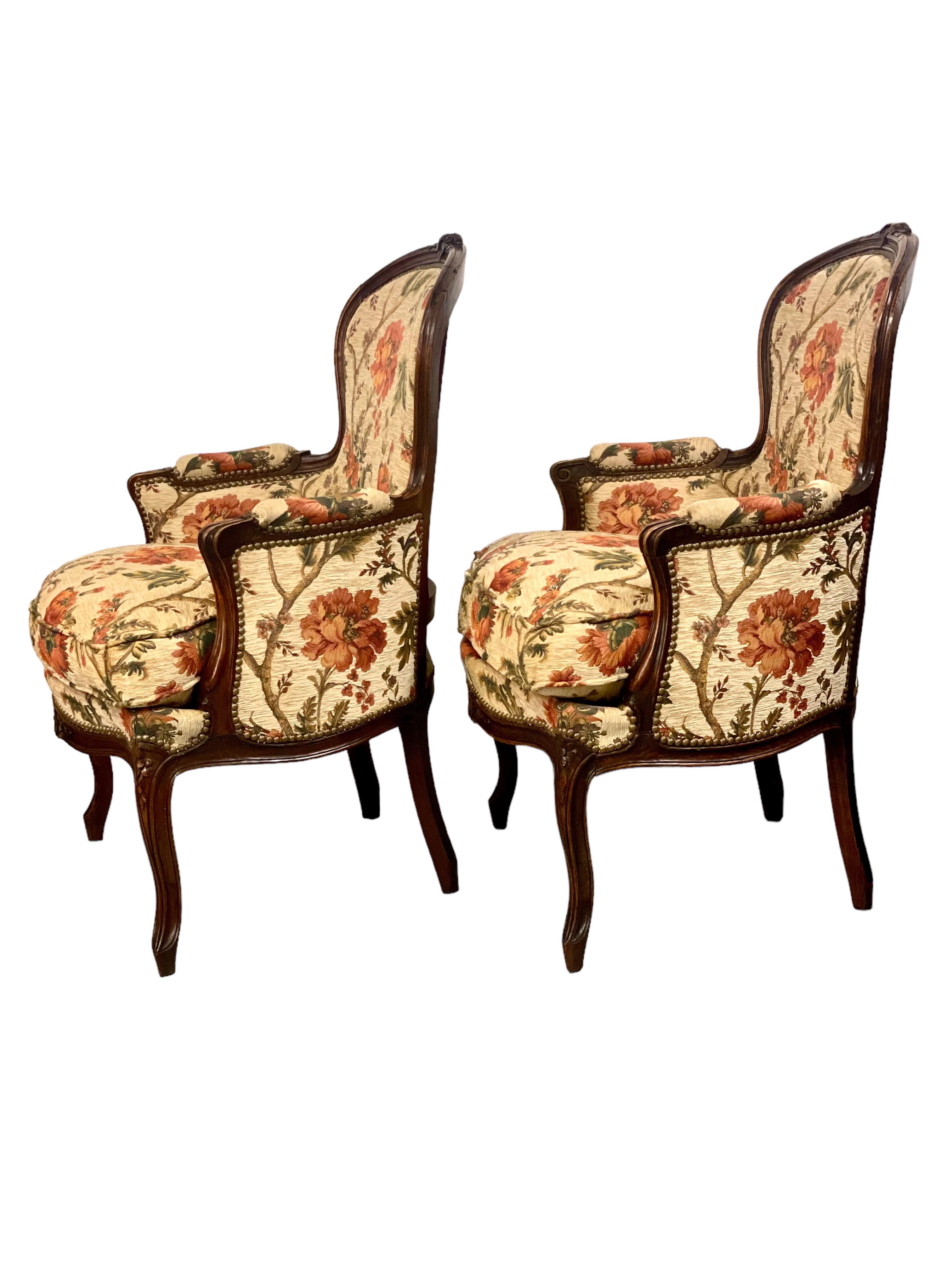 A matching pair of elegant and extremely comfortable Louis XV style 'Bergère' armchairs, upholstered in a luxurious woven floral fabric in warm shades of apricot, terracotta and green set against a cream background. 
The curving wooden frames are
