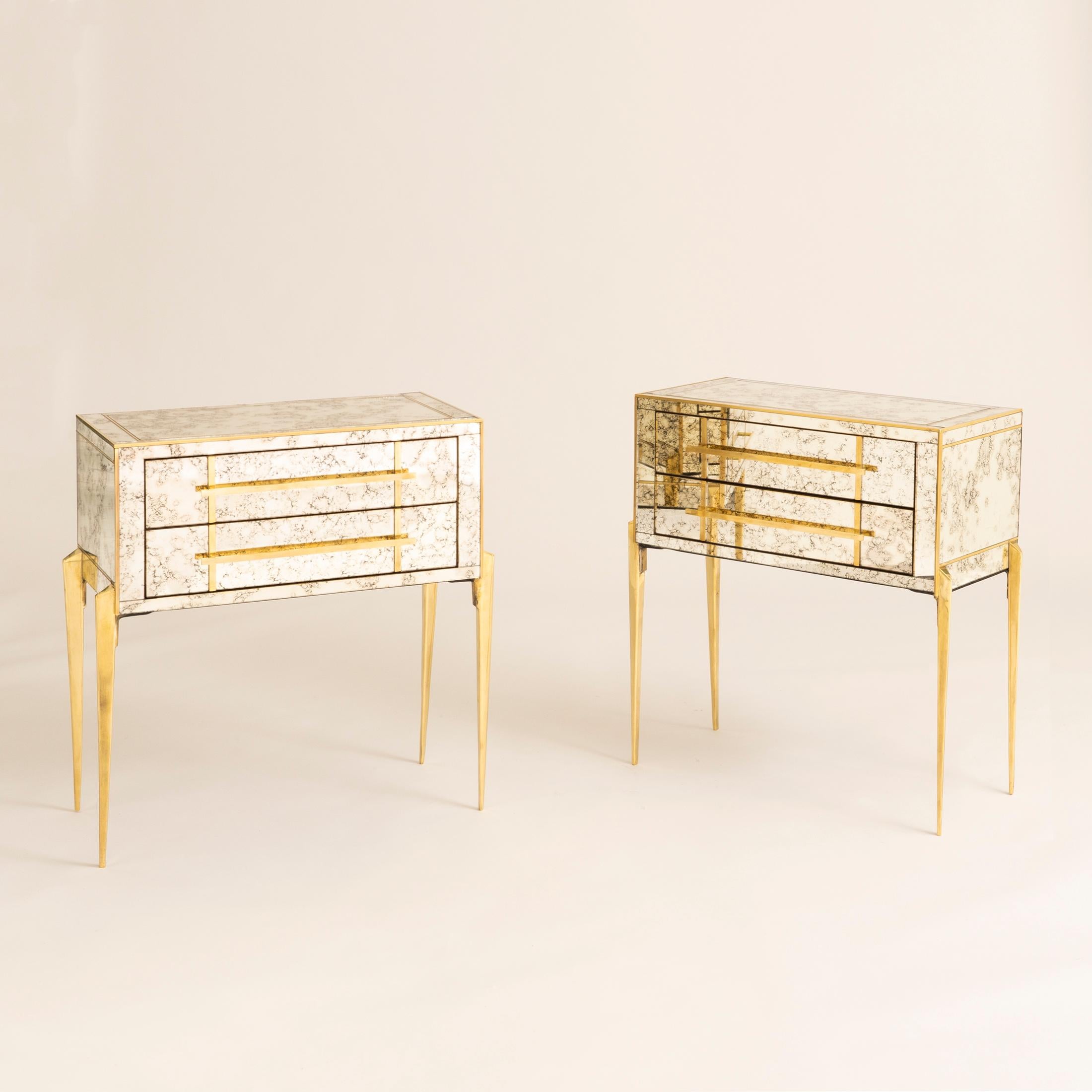 Pair of unusual wide two-drawer chest of drawers/bedsides in antique mirror surrounded by brass trim. Sits on triangular shaped finely tapered brass legs positioned on the outside of the tables. A glamorous addition to any room.