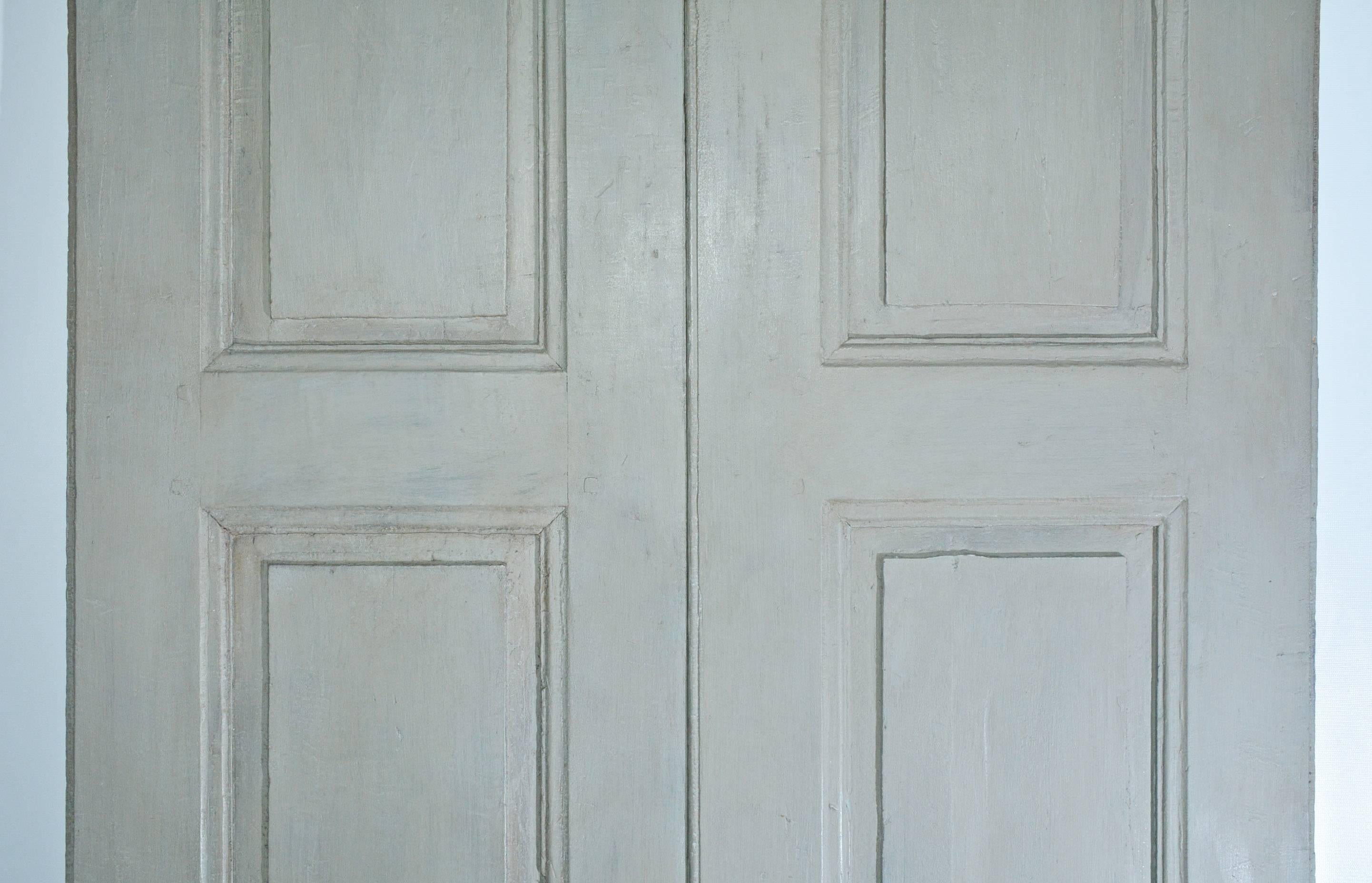 The pair of French antique doors or shutters are painted grey. Each door has two long panels over one short panel. The backsides have two-over-one simpler panelling. There is a key hole and half of the two pairs of hinges.