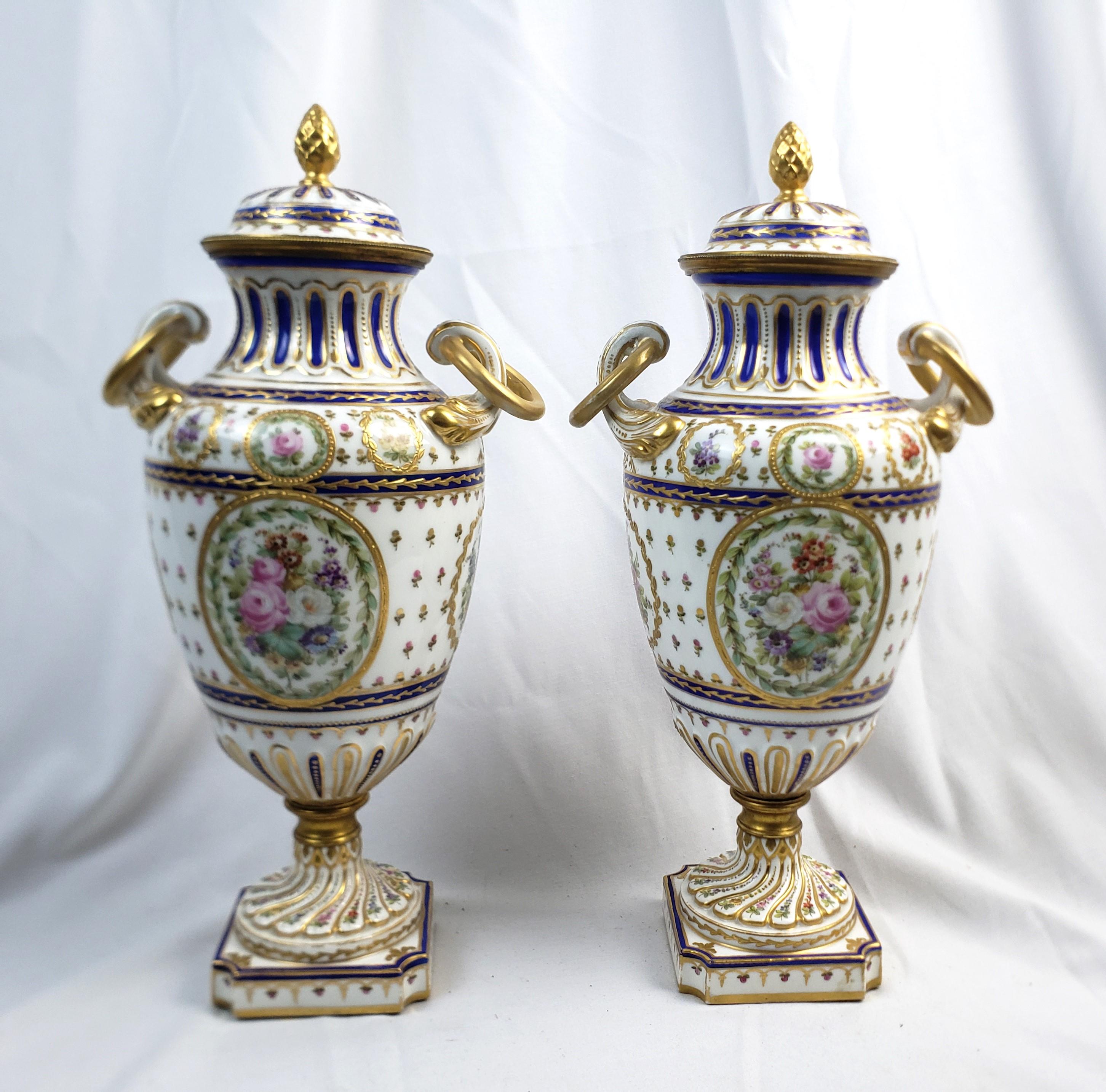 This pair of antique covered urns are signed by and unknown maker, and presumed to have originated from France and date to approximately 1880 and done in the Sevres style. The covered urns are done in porcelain with a white grouns and cobalt blue