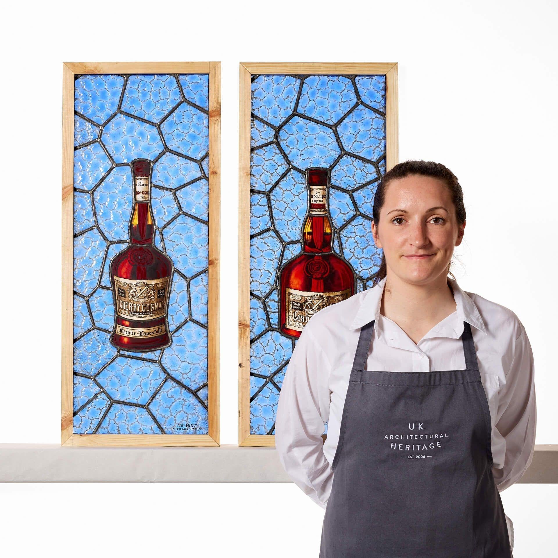 Paire de vitraux anciens de liqueur signés par Mon Giot, Vitraux, Paris. Cette paire de panneaux de vitrail présente des images vives et lumineuses de deux bouteilles de liqueur, un cognac à la cerise et un grand marnier, marquées 1827. Les deux