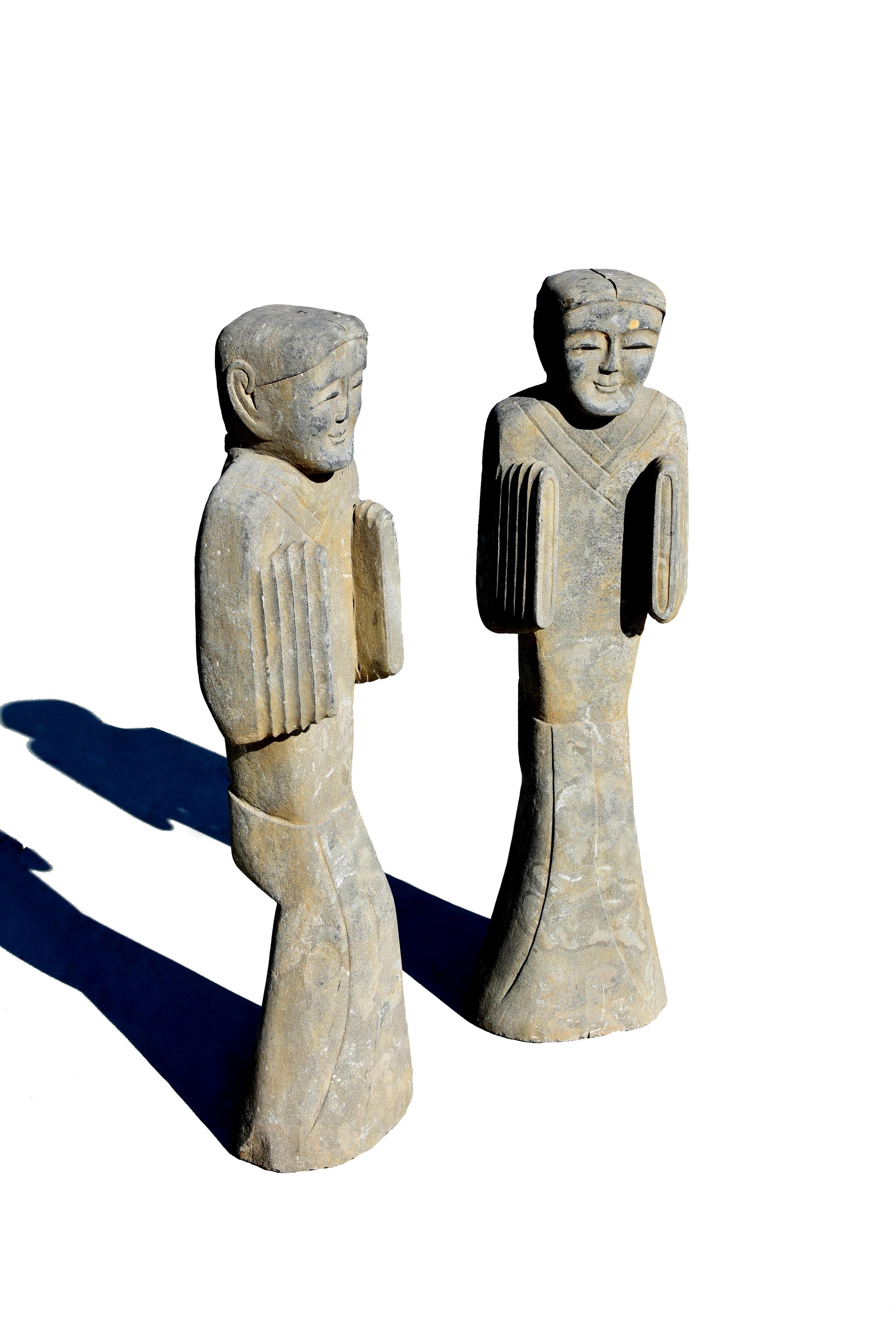 A pair of vintage solid stone court attendants. Of tall slender form, wearing long flowing robes flaring at the bottom with hands within wide, decorative cuffs. The realistically modelled face with serene expression is with large, kind eyes under