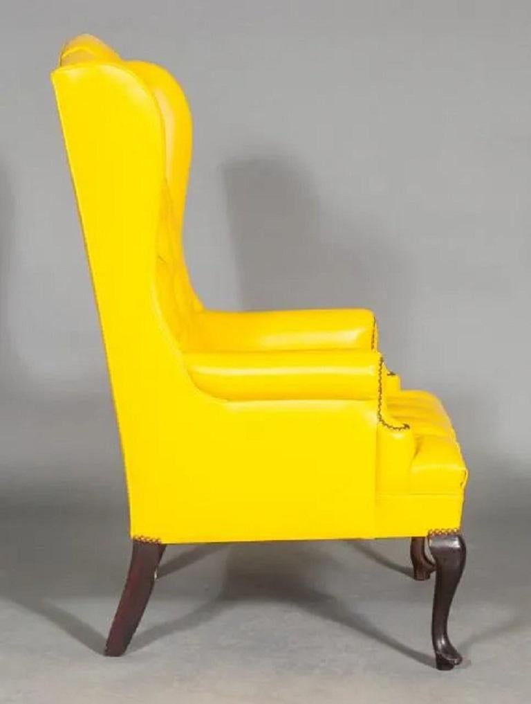 Pair of antique style wingback chairs. The chairs have tufted backs and seats, with yellow leatherette upholstery, brass tacks to the front of the arms, and solid wood cabriole legs.