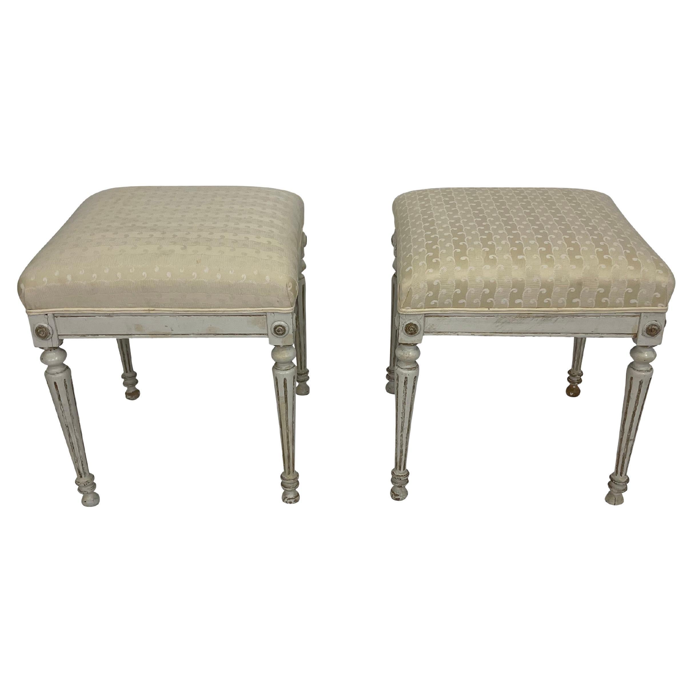 Pair of Swedish Gustavian Ottomans in white paint and parcel gilt. In a white painted finish, the carved frames in the Louis XVI style with cylindrical fluted tapering legs and toupie feet. These stools have been professionally restored to their